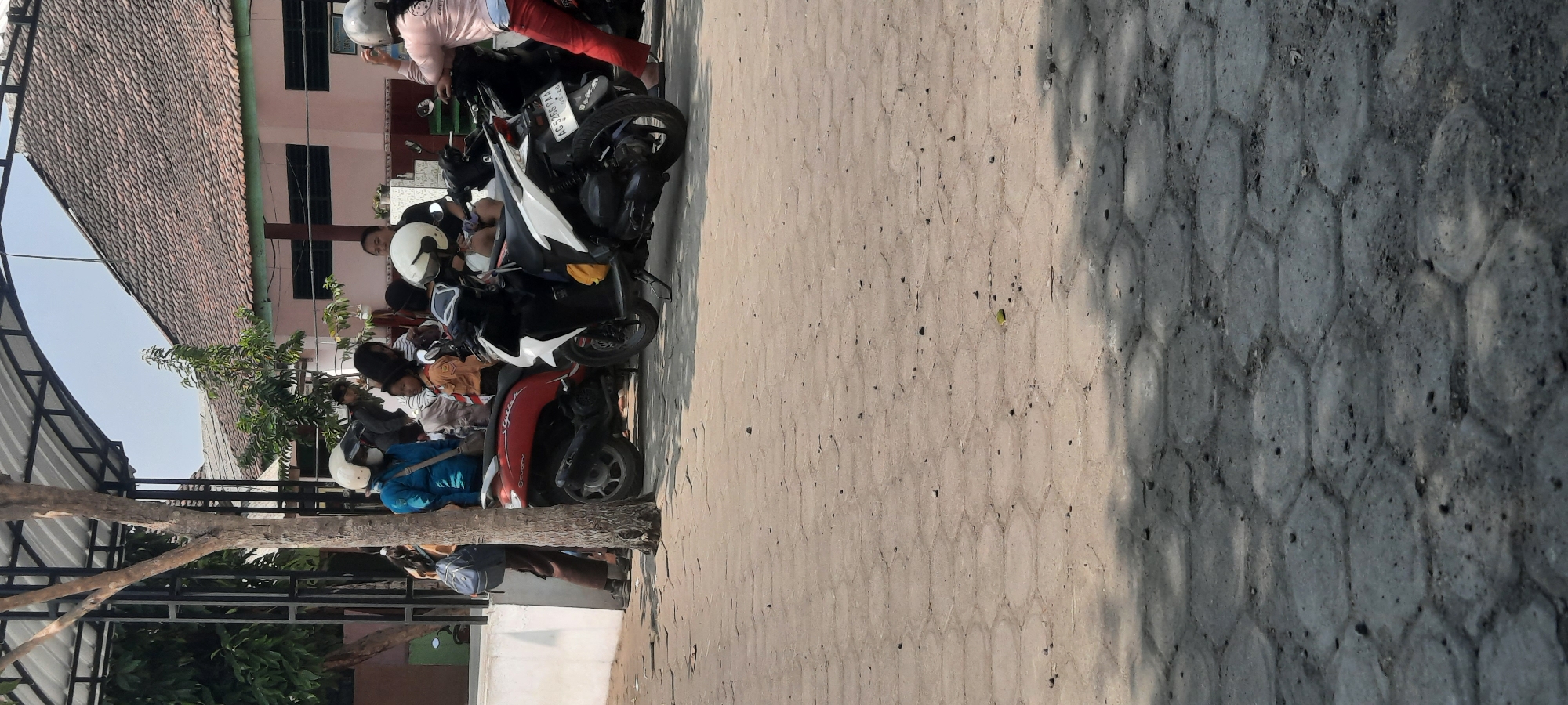 In Tangkil, Indonesia, several individuals are gathered near parked motorcycles under a covered structure. On the left, a person in a blue jacket and white helmet stands beside a red scooter, holding a blue backpack. To their right, a group of people are milling around two other motorcycles, one white and one black. The white motorcycle has a yellow bag on its seat and a license plate that reads "AG 5266 PAA". In the background, a building with a tiled roof and pink walls is visible. The ground is paved with hexagonal stones, and tree shadows are cast across it. The scene appears to be an outdoor waiting area or gathering point, possibly near a school or public facility.
