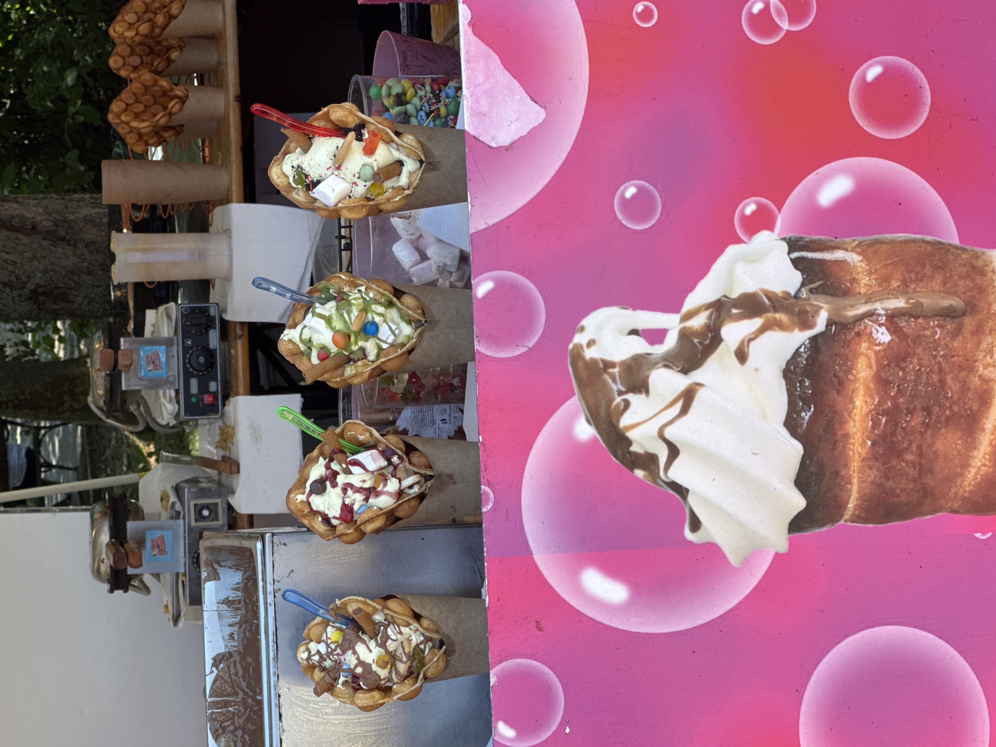 Four bubble waffle ice cream cones with various toppings are displayed on a counter. In the background, a waffle maker and containers of ingredients are visible. The counter is positioned in front of a pink backdrop with illustrations of bubbles. The scene appears to be at an outdoor food stall in Burgas, Bulgaria, likely during daylight hours. No people are visible in the frame.