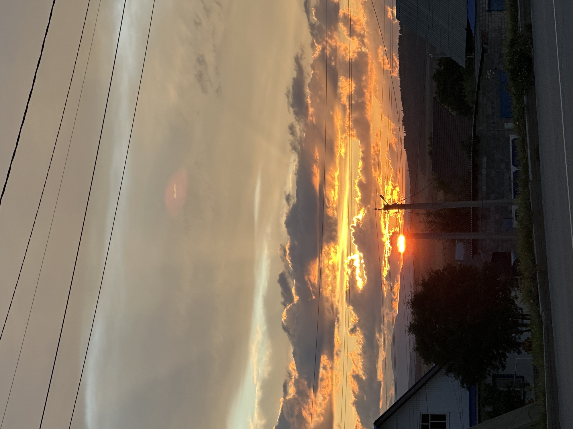 The image depicts a dramatic sunset with vibrant orange and yellow clouds illuminating the sky. The sun is partially obscured by clouds, casting a warm glow over the scene. In the foreground, several power lines stretch diagonally across the frame, creating a sense of depth and urban influence on the natural landscape. A utility pole stands to the right, supporting the wires.
The background features a dark, silhouetted mountain range, providing a stark contrast to the luminous sky. Below the mountains, hints of human habitation are visible, including the rooftops of houses and buildings, suggesting a rural or semi-urban setting. A dark tree stands out prominently, its branches silhouetted against the colorful sky. A portion of a road is visible on the far right, indicating it is a roadside view. The lighting suggests that the photo was taken during the golden hour, just before sunset. The overall mood is one of awe and natural beauty, with a touch of human presence. There is no visible text in the image.