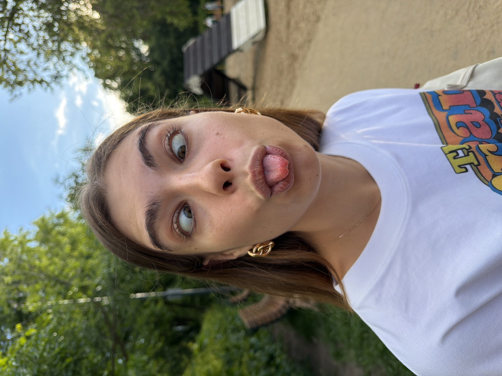 A young woman stands outdoors with trees and a building in the background. She is wearing gold chain earrings and a white t-shirt with a colorful design. The woman is sticking out her tongue and crossing her eyes. It is daytime. Given the type of architecture and vegetation visible, the location is likely Moscow, Russia.