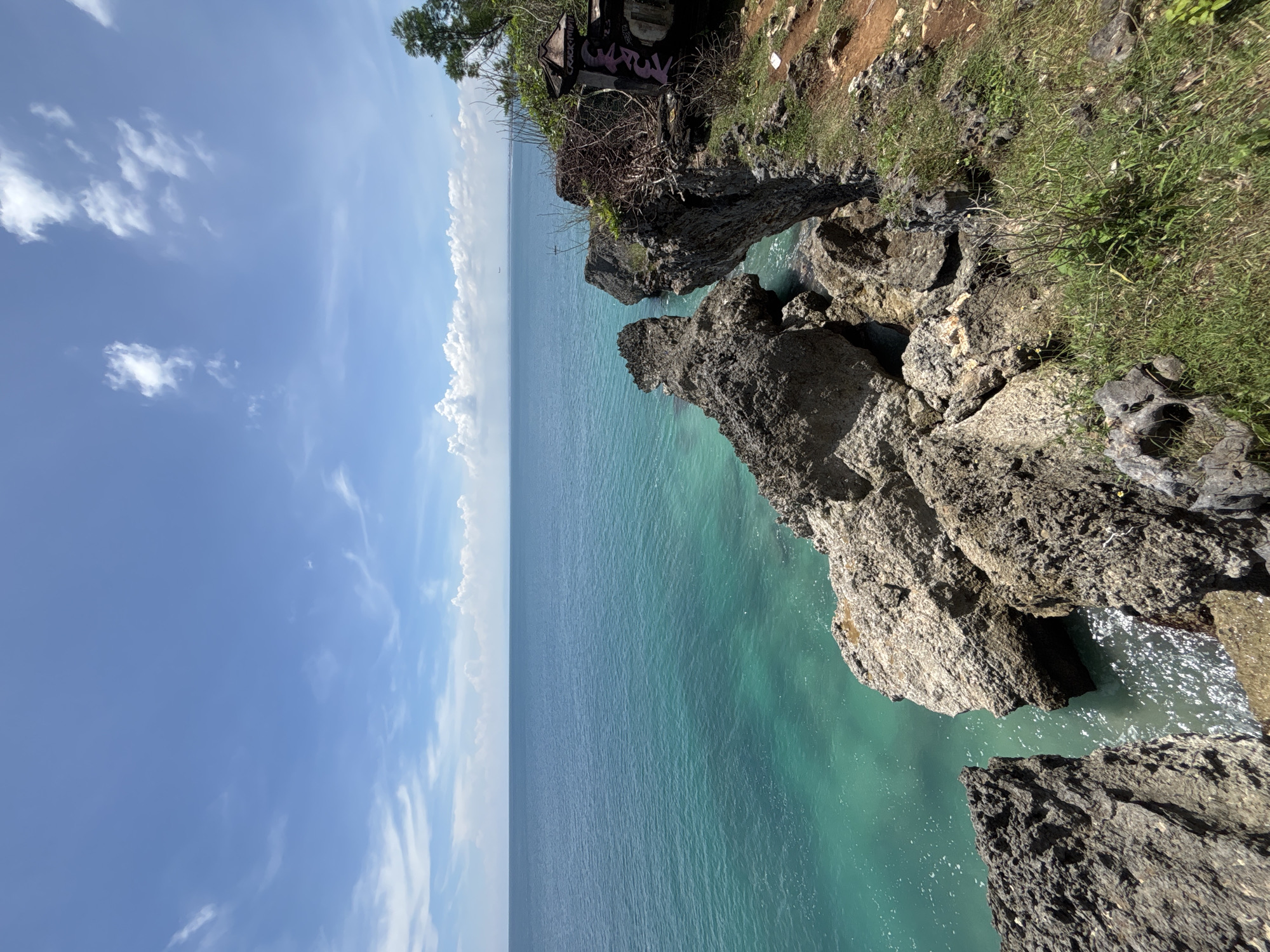 The image captures a coastal scene in an area with rocky cliffs, likely in or near Unknown, Indonesia. The scene is composed of the sky, ocean, and rocky cliffs, which dominate the right side of the frame. The sky is light blue with scattered clouds, transitioning into the turquoise color of the ocean. The ocean stretches out to the horizon, with a clear separation between the sky and the sea. The rocky cliffs are covered in vegetation, including grass and some trees. The rocks are gray, with rough texture. The water near the cliffs is clear, revealing the rocky underwater landscape. There are no people visible in the image.