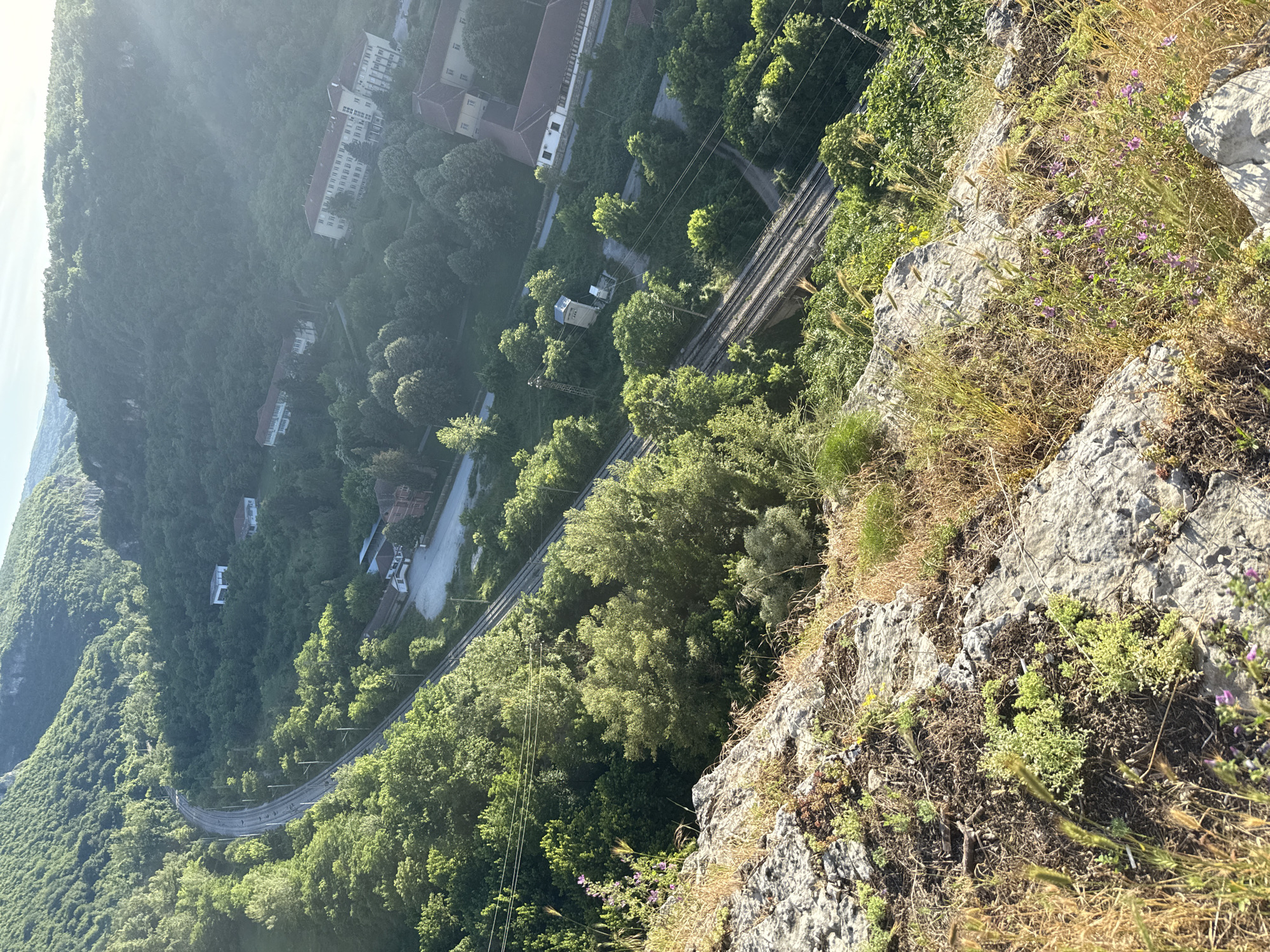 The image is a high-angle view looking down into a valley in Karlukovo, Bulgaria. The foreground features a rocky ledge with dry grasses and small purple flowers. Below, a train track curves through a dense, green forest. Several buildings are visible nestled within the trees, including what appears to be a residential area or institution with multiple structures. The sun is shining, casting a bright light and lens flare on the right side of the image, highlighting the lush vegetation and the sprawling buildings. A road is also visible running parallel to the train tracks in some sections. The overall scene depicts a natural landscape integrated with human infrastructure.