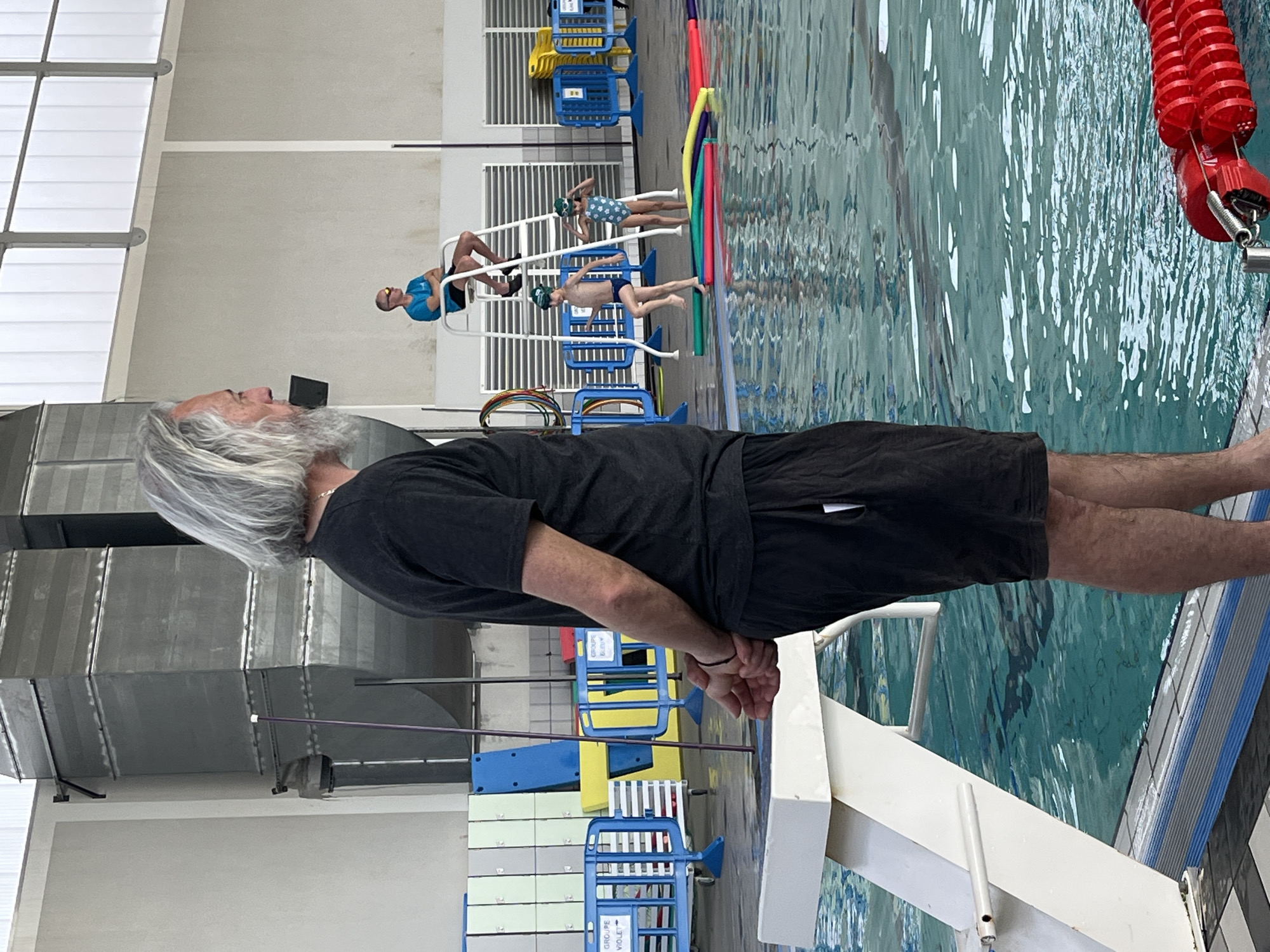 The image depicts the aquatic center in Andrézieux-Bouthéon, France. An older man with grey hair and beard, wearing a black t-shirt and shorts, stands near the edge of a swimming pool. In the background, several children are lined up on starting blocks, apparently preparing to swim. Red lane dividers are visible in the pool. The environment is brightly lit.