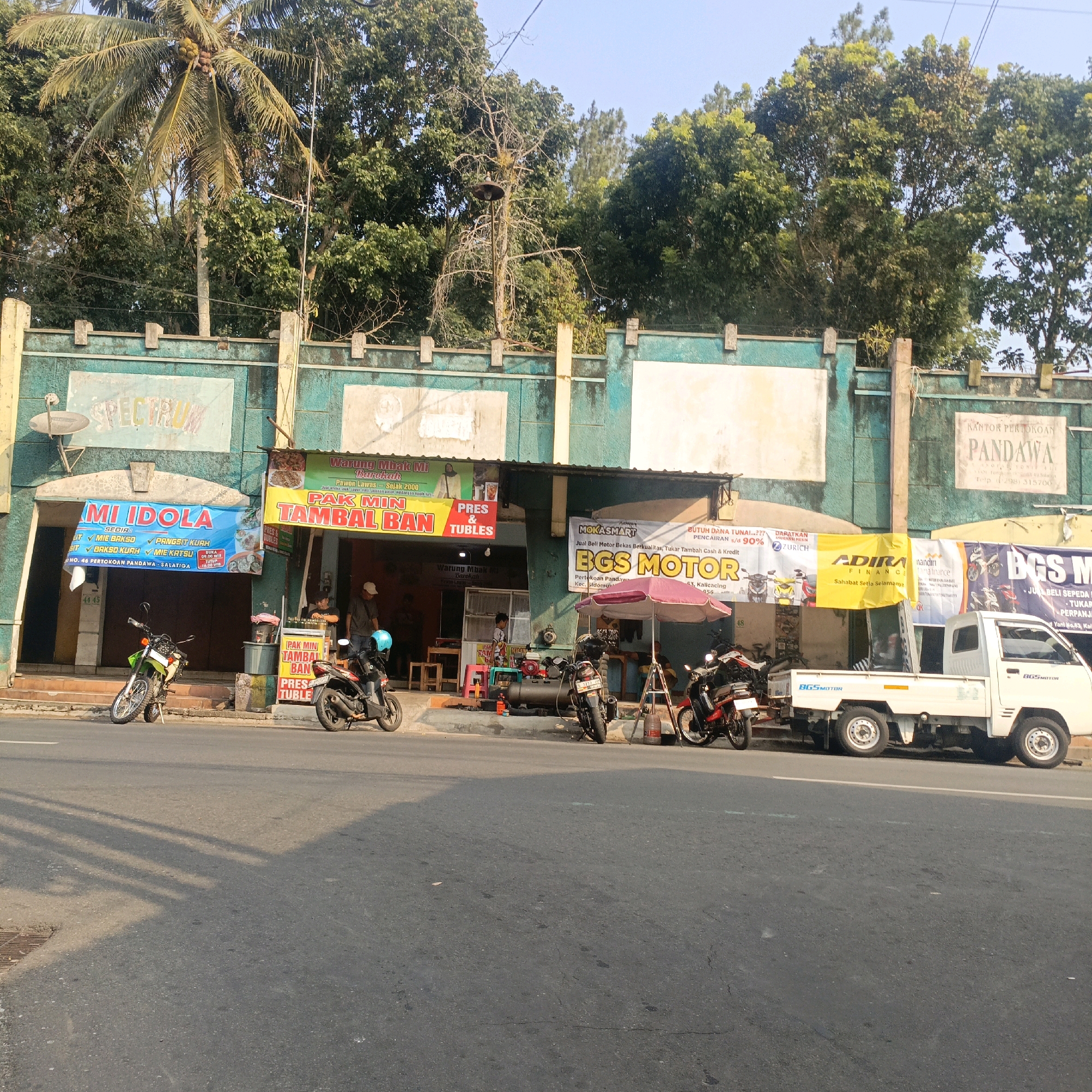 The image depicts a street scene in Salatiga, Indonesia, in front of a row of businesses. The businesses appear to be a mix of food stalls and vehicle repair shops, indicated by signage such as "Warung Mbak Mi Barekah" (Mrs. Mi's Blessing Stall) and "BGS MOTOR" (BGS Motor). Several motorcycles are parked along the sidewalk in front of the shops, and a white pickup truck is parked on the street. The architecture of the buildings is characterized by a green facade with white accents. Trees line the background, suggesting a somewhat green urban environment. The lighting indicates it is daytime, with shadows cast on the road. There are at least two people visible, one standing near a motorcycle and another partially visible inside one of the shops. A satellite dish is mounted on the wall of one building, and various banners and advertisements are displayed.
