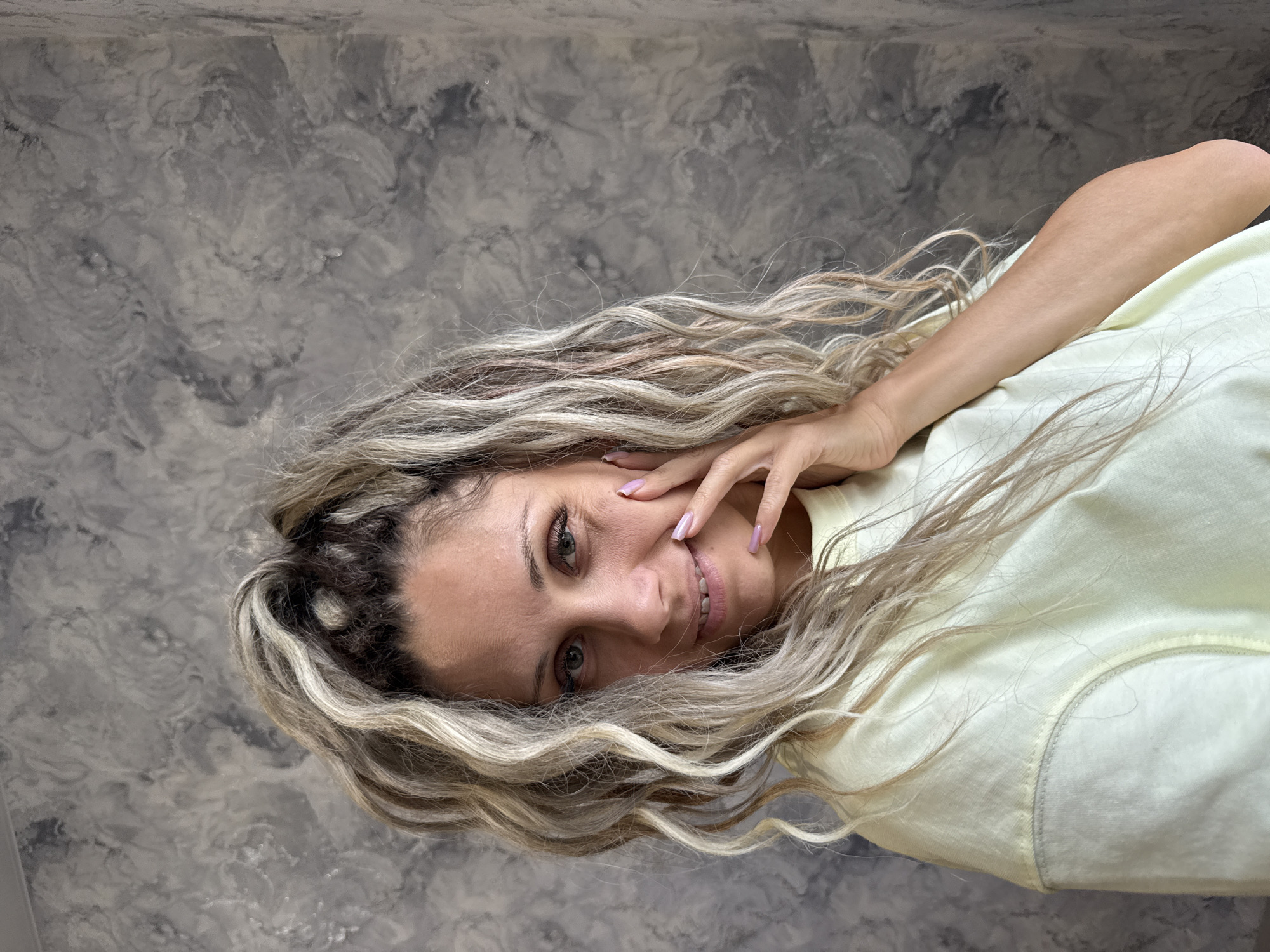 A woman with blonde dreadlocks is positioned in the center of the frame, facing the camera with a slight smile. Her right hand is raised to her face, with her fingers gently touching her cheek. She is wearing a light yellow, crew-neck sweatshirt. The background is a textured gray wall. The image was taken in Rusanovo, Russia.