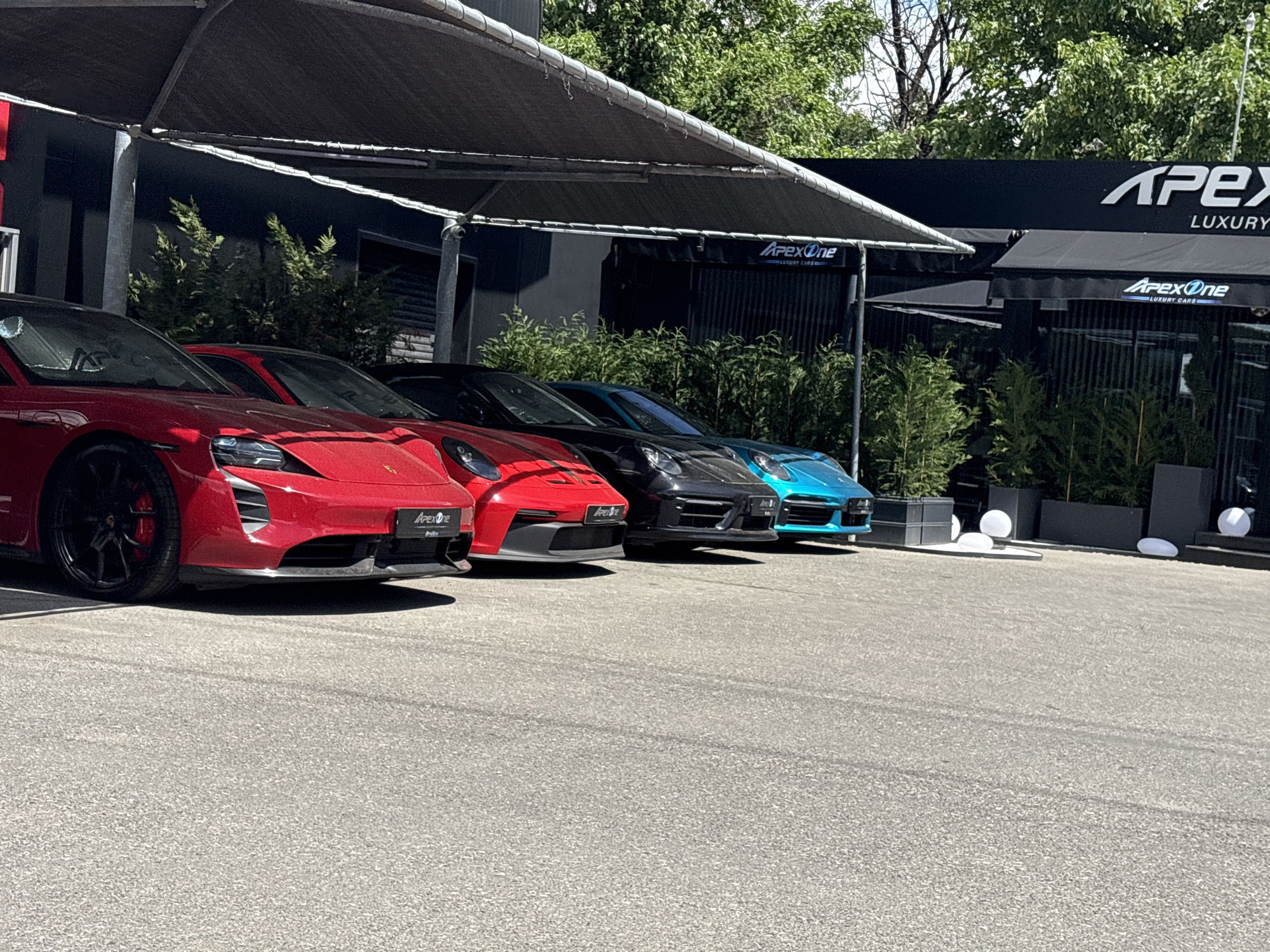 The image shows a row of four Porsche cars parked in front of a building with "Apex Luxury Cars" signage. The cars are in the following colors from left to right: red, red, black, and turquoise. A dark awning covers the parking area. The building is modern with dark finishes and landscaping. The location is likely in Sofia, Bulgaria. The parking area is paved and looks to be freshly swept. There are white decorative orbs on the sidewalk near the building. No people are visible in the frame.