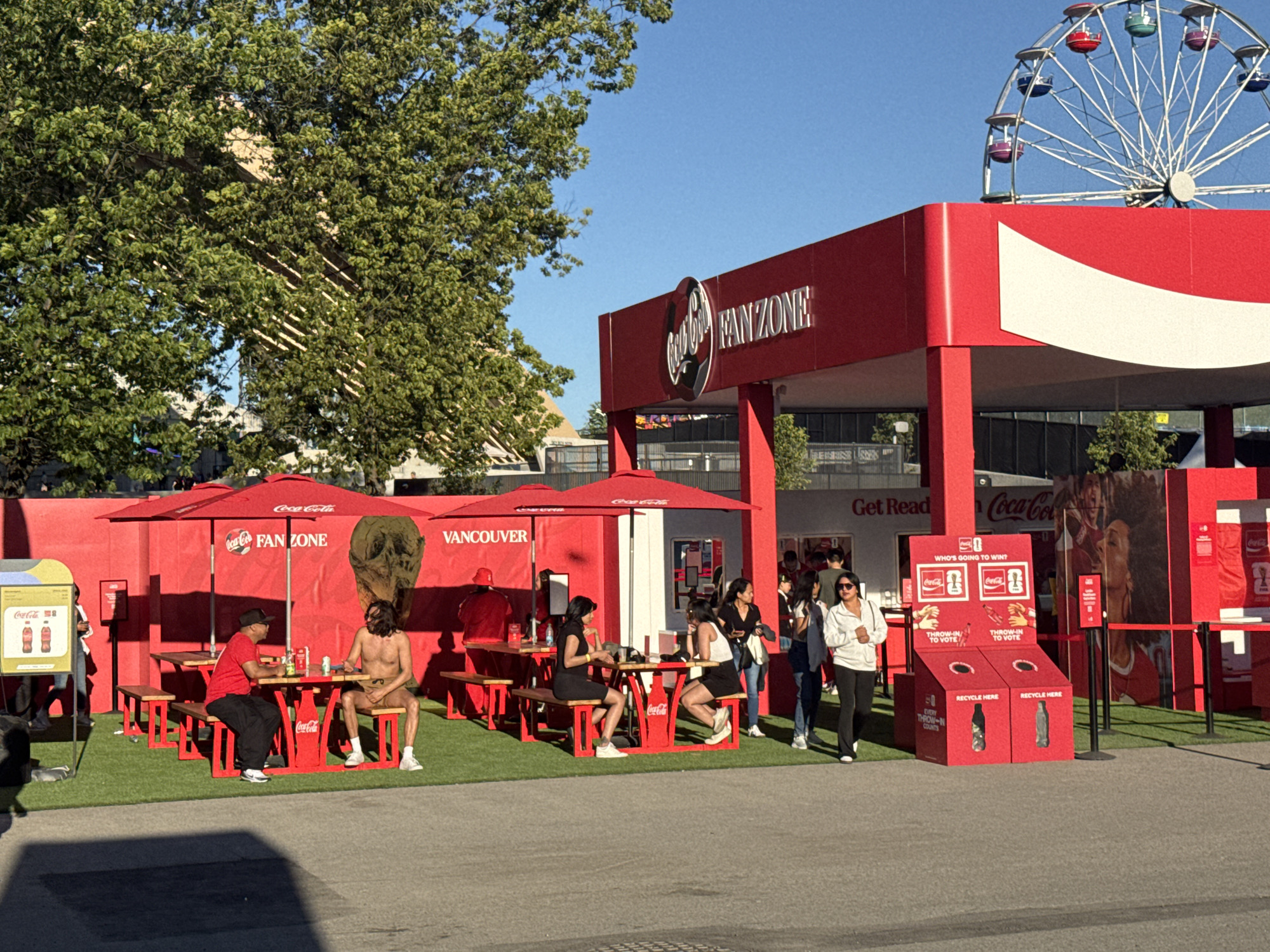 The image shows an outdoor Coca-Cola Fan Zone in Vancouver, Canada, likely set up for a sporting event. A large red structure with "Coca-Cola FANZONE" signage is prominent. In the foreground, several picnic tables with red umbrellas are set up on artificial turf. People are seated at the tables, some interacting, others appearing to rest or observe. One individual is seated shirtless at a table. In the background, a Ferris wheel is visible against a clear blue sky, suggesting a public event or amusement park setting. Red recycling bins are placed near the structure, and promotional posters are displayed on the walls. The overall scene depicts a vibrant, branded promotional space.