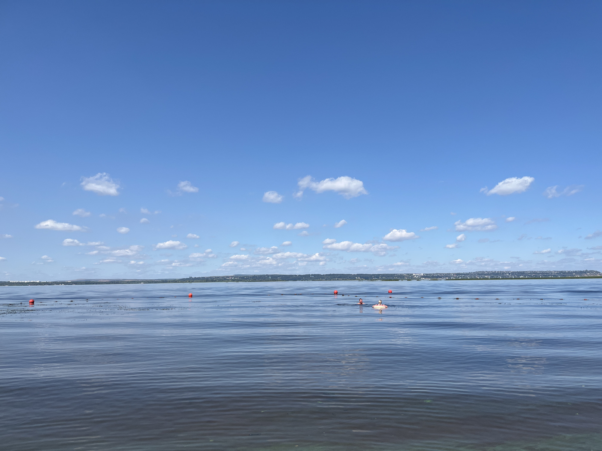 A bright, sunny day is depicted over a calm body of water with a distant shoreline. The sky is a vibrant blue with scattered white cumulus clouds. The water's surface exhibits gentle ripples. In the middle distance, a line of orange buoys is visible, demarcating an area on the water. Two individuals are present in the water, swimming amongst the buoys. One person is further to the left, their head and shoulders visible above the surface, wearing a red cap. The other person is closer to the right, partially submerged and appears to be using a yellow inflatable ring. The distant shoreline is lined with trees and buildings, suggesting a populated area. The location is identified as the city of Shumeika, Russia.