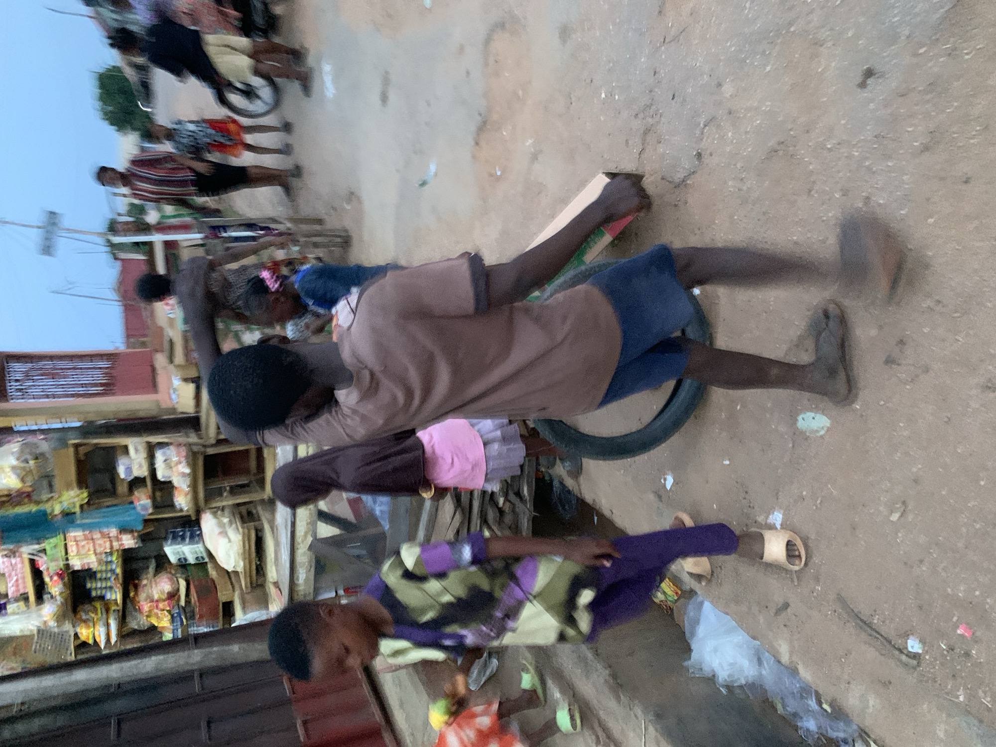 This blurry image captures a bustling street or market scene in Kabba, Nigeria, likely during dusk or dawn, as indicated by the soft, diffused light and a pale blue-grey sky. The ground is unpaved and dusty, with scattered debris including plastic bags.
The foreground and mid-ground are populated by several people, mainly children. A young person, centrally positioned, wears a brown short-sleeved shirt and blue shorts. This individual holds a wooden stick or plank in their right hand, appearing to interact with or push a tire lying flat on the ground, possibly as part of a game or activity. Their bare right foot and a light-colored sandal on the left foot are visible. To the left, another child, clad in a purple and green patterned shirt and purple trousers with light-colored sandals, stands and looks towards the main activity. Further back, another person wears a pink skirt.
On the left side of the frame, several wooden market stalls or shelves are packed with various packaged goods, suggesting an active street market. In the blurred background, other figures, including one on a bicycle or motorcycle, contribute to the sense of movement and activity in the street. The overall blur indicates motion, either from the subjects or the camera. No discernible text is visible on any signs or products due to the blur and distance.