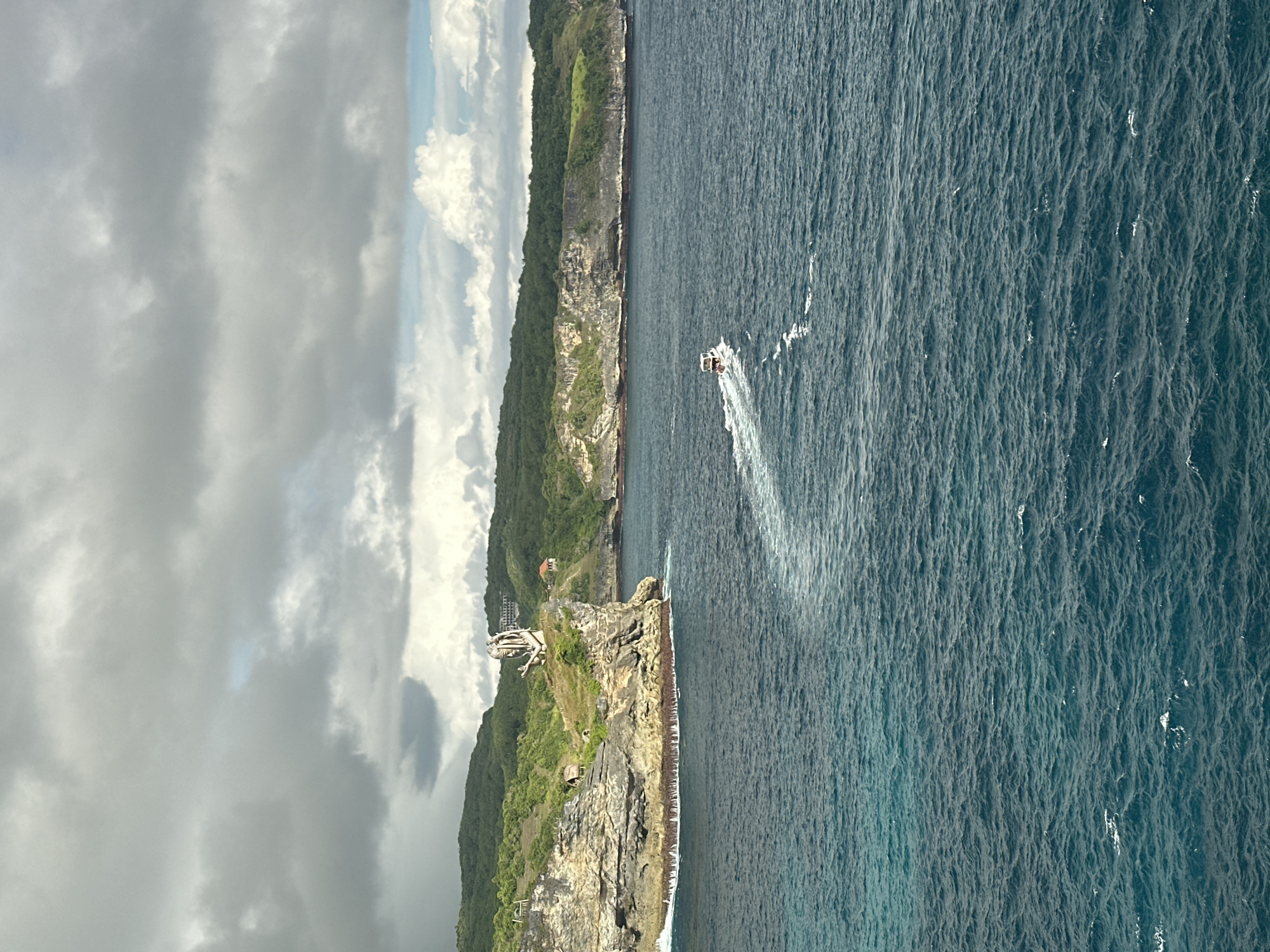 A full shot reveals a coastal view in Nusa Penida, Indonesia, under a partly cloudy sky. The scene is defined by a rugged coastline, densely covered with green vegetation, contrasting with the deep blue of the ocean. A jet ski is observed moving through the water, creating a white wake. On the cliff, a structure is visible.
