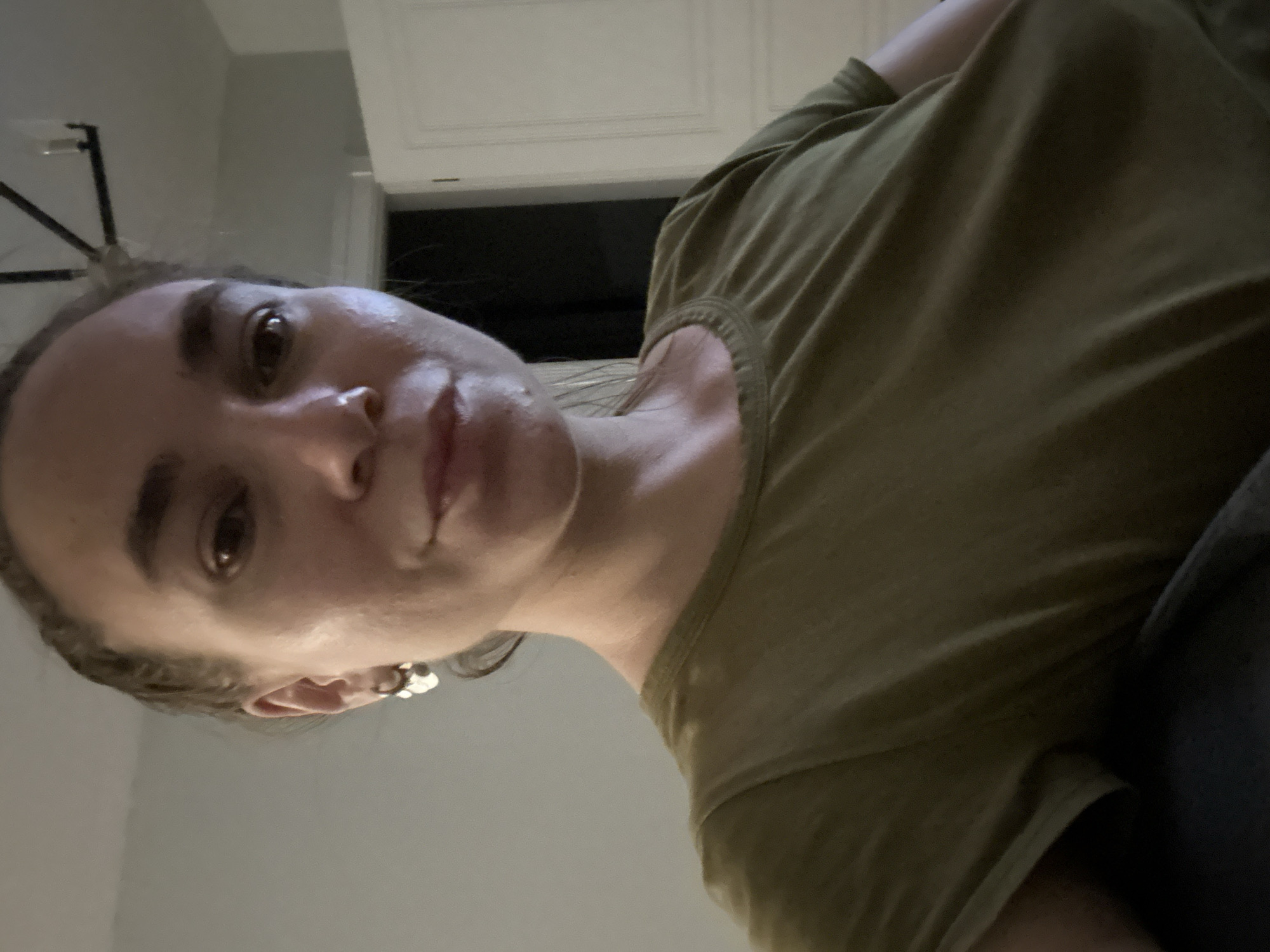 The image shows a close-up, low-angle view of a person with light skin, brown hair, and brown eyes, wearing a green t-shirt. The person is looking directly at the camera. The background appears to be an interior space, with a white cabinet or door visible in the upper portion of the image. A dark area is visible behind the cabinet. A light fixture can be seen in the upper left corner of the image. The person is wearing earrings.
There is no information in this image to indicate the location is in the city of Kharnavy, Russia.