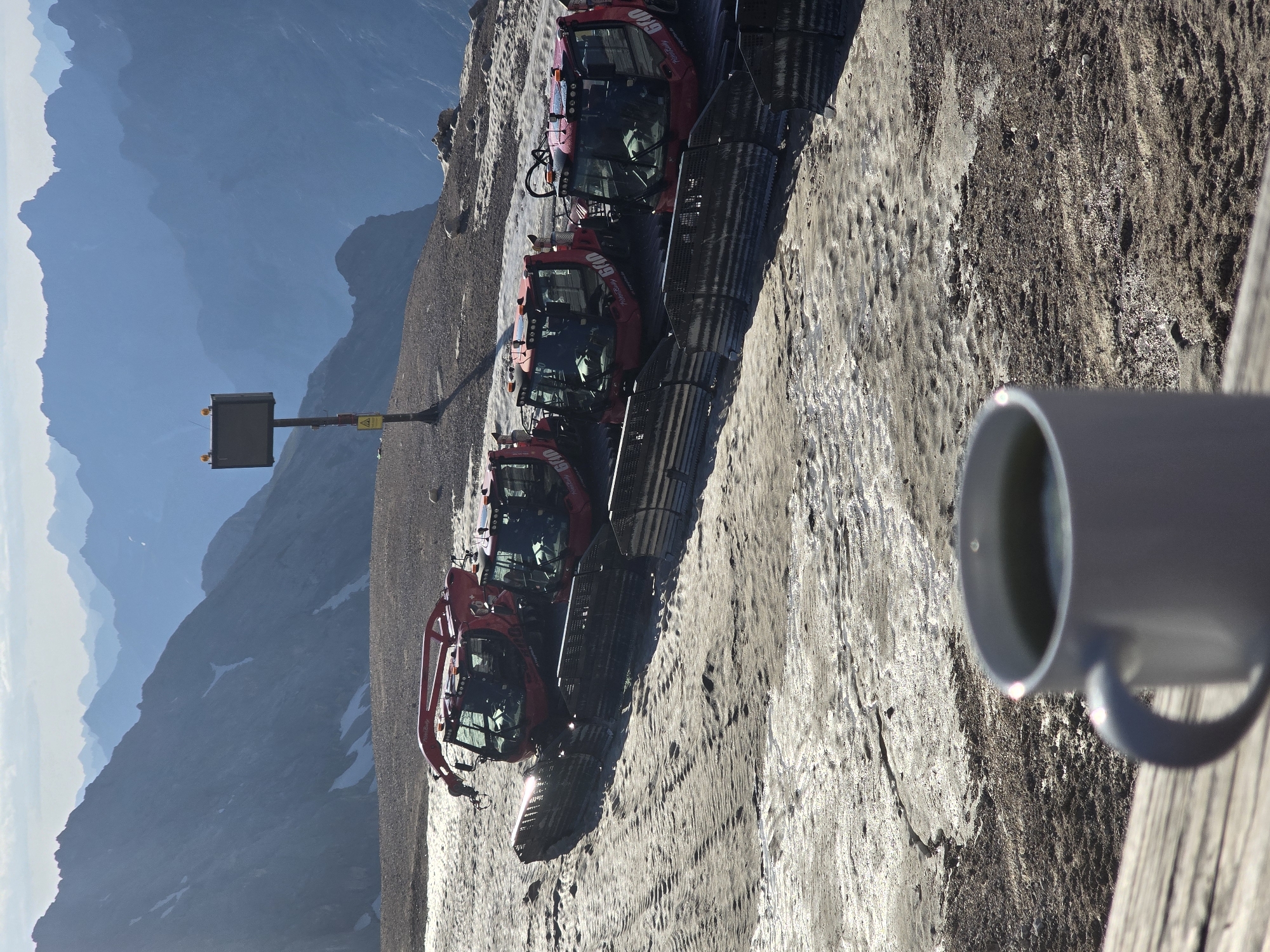 The image captures a scene at the Zugspitze, Germany's highest peak, near Garmisch-Partenkirchen. In the foreground, a white mug filled with a green beverage sits on a wooden surface. Beyond the mug, several red snow groomers are parked in a line on a sandy and icy terrain. The snow groomers are positioned behind a black metal barrier. In the background, a series of majestic blue mountains stretch across the horizon under a bright sky. A tall pole with a rectangular sign and warning symbols stands on the right side of the frame. The overall environment suggests a cold, mountainous region.