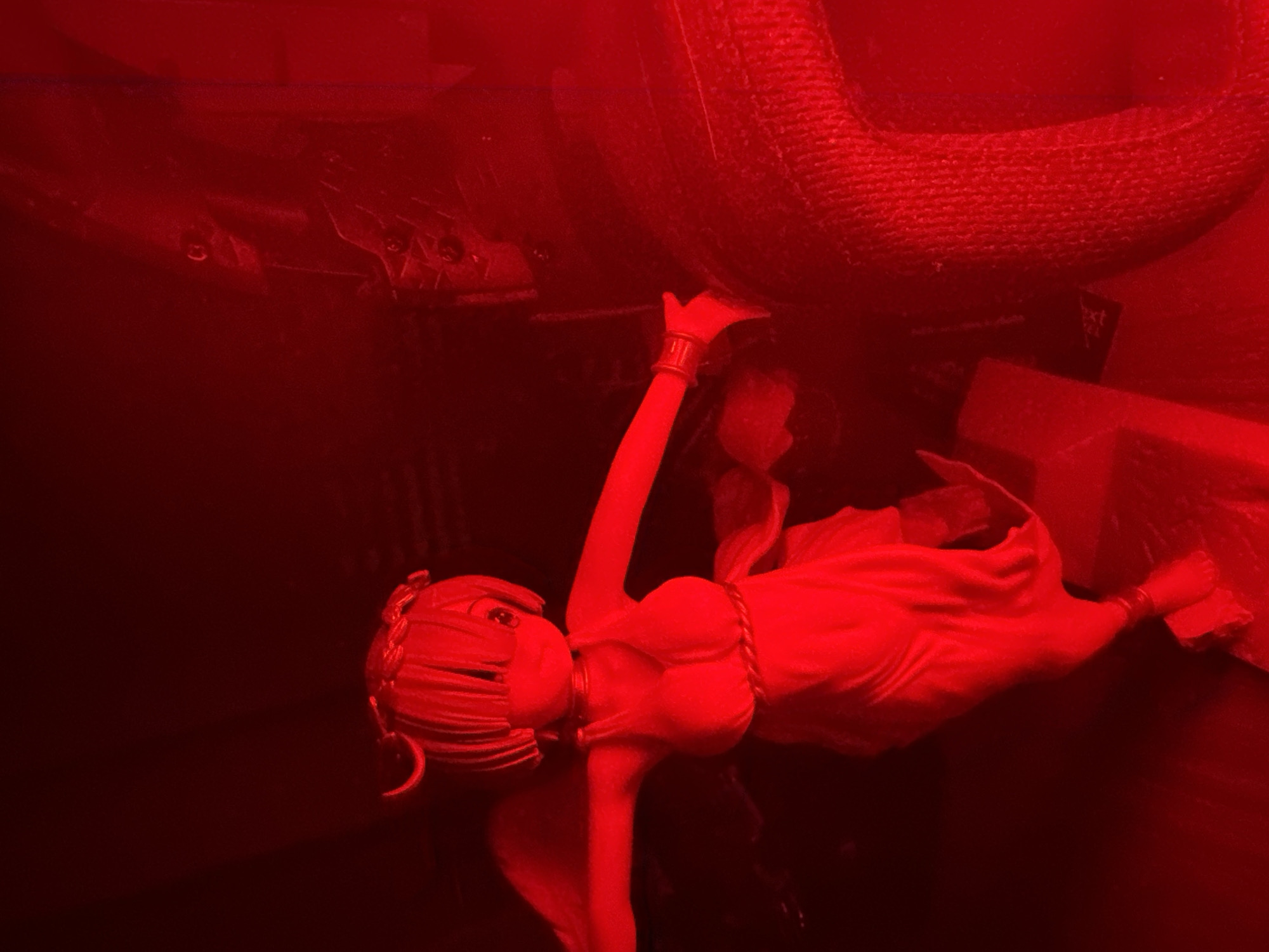 An anime figurine of a female character with light-colored hair and a red halo is positioned in the center of the frame, bathed in red light. The figurine is wearing a white dress with a gold belt and appears to have small wings. It is posed with arms outstretched. To the right of the figurine, a large, textured object, possibly a piece of furniture or equipment, is visible, also illuminated by the red light. The background is dark and indistinct, with some shadowy shapes suggesting an indoor environment. The overall impression is one of a staged display or collection illuminated by a red light source.