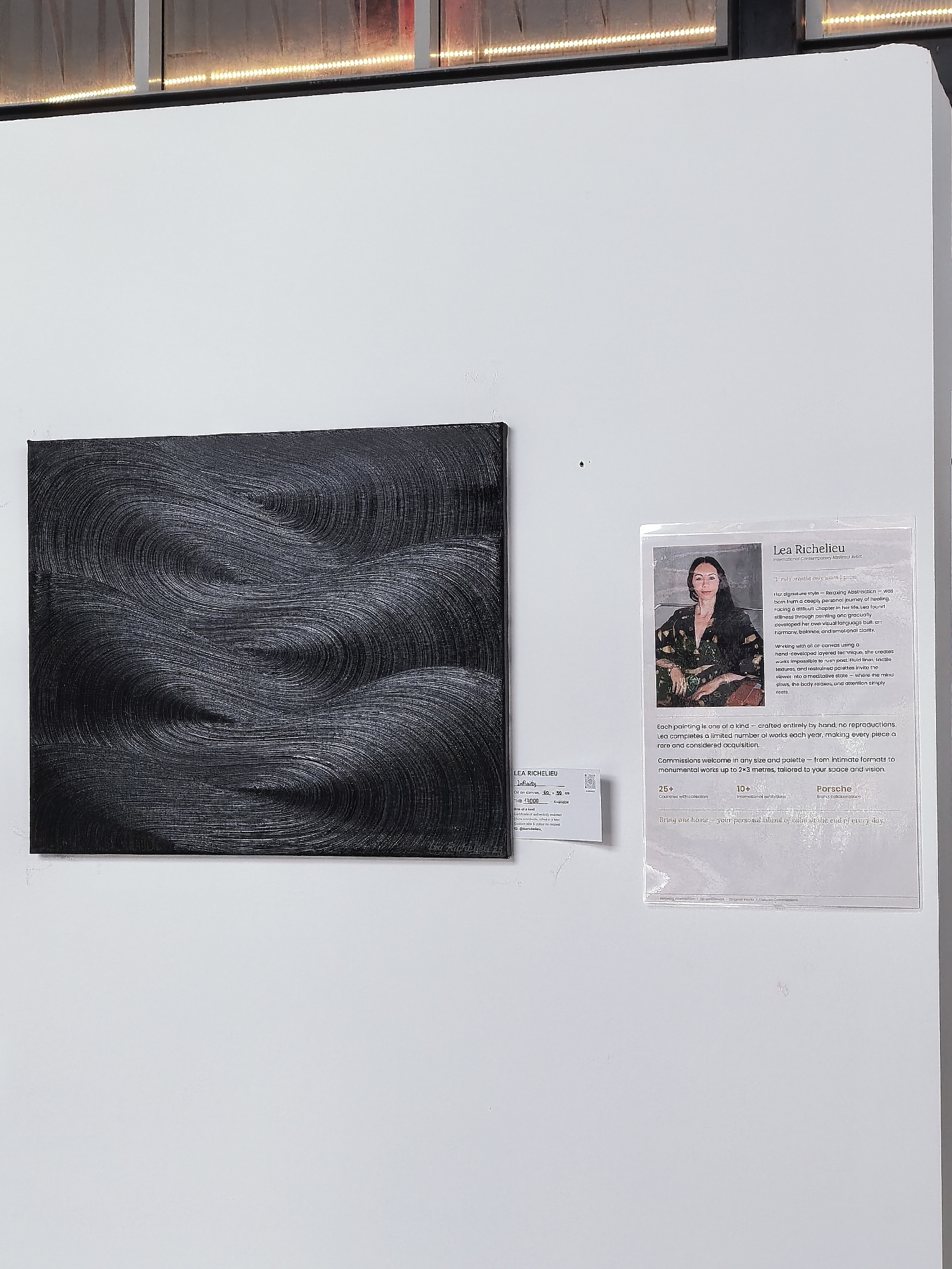 The image depicts an art exhibition in a gallery space. On the left is a black abstract painting with textured, swirling patterns. To the right of the painting is a white wall with two pieces of information. The first is a small, white card with text that appears to be artwork details, including the artist's name "LEA RICHELIEU," the artwork title "Infinity," dimensions "60 x 80 cm," and price "THB 40000." The second is a larger, informational placard featuring a portrait of the artist, Lea Richelieu, and biographical and artistic statements. The placard includes her name prominently at the top. The background of the gallery shows a wall and what appears to be an illuminated ceiling structure. There is no visible indication that this scene is located in Ko Samui District, Thailand. The description of the scene is forensic, matter-of-fact, and includes essential details about the subjects, objects, and environment.