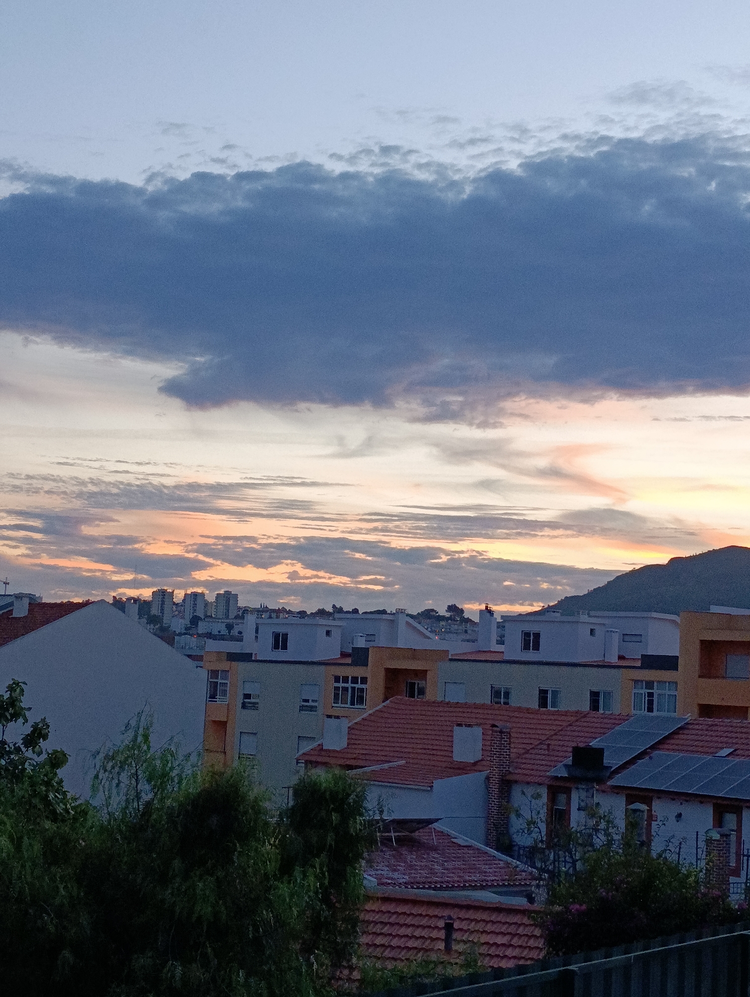 This image captures a dusk scene over a city, likely Setúbal, Portugal, viewed from an elevated position. The sky is dominated by a gradient of colors, transitioning from a pale blue at the top to warm orange and pink hues near the horizon, indicative of sunset. Dark, textured clouds are present in the upper portion of the sky.
Below the sky, a cityscape unfolds. Buildings of varying heights and architectural styles are visible. In the middle ground, several taller apartment buildings with multiple windows are clustered together. In the foreground and mid-ground, smaller residential buildings with red-tiled roofs are prominent. Some of these buildings feature solar panels on their rooftops. Trees with dark green foliage are scattered throughout the scene, some in the foreground obscuring parts of the buildings. A dark fence runs along the bottom edge of the image. The overall impression is a quiet urban environment at the end of the day. There are no people or animals visible in the image.
