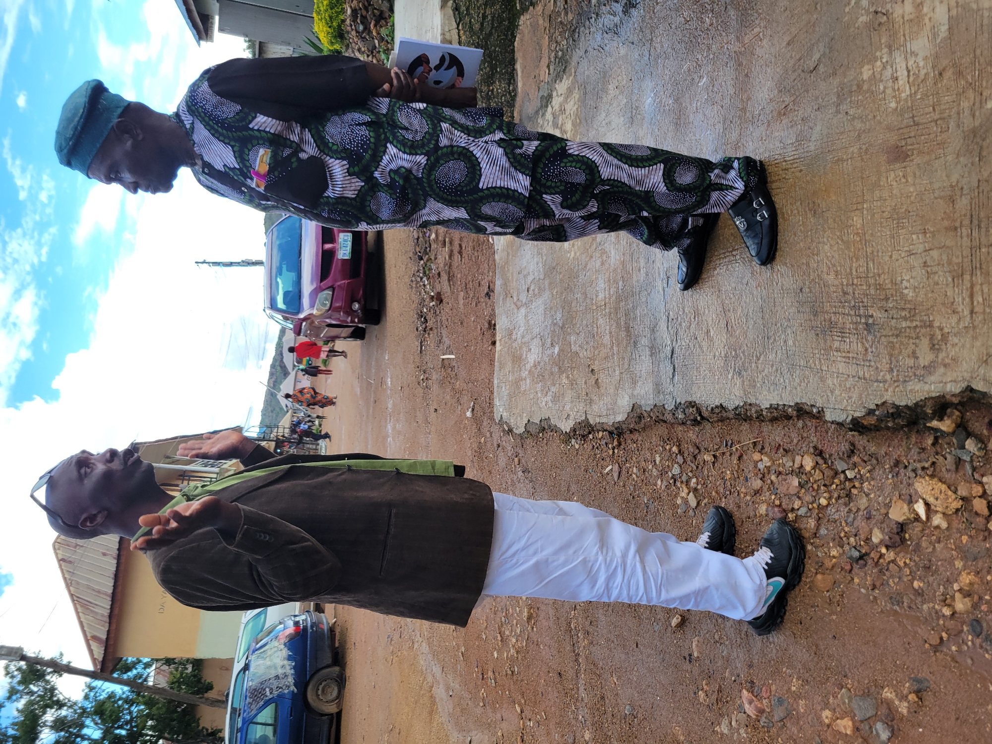 Two men are standing and talking in a street in Jos, Nigeria. The man on the left is wearing a brown jacket over a green shirt and white pants, with black and blue sneakers. The man on the right is wearing a green and white patterned traditional African outfit and black shoes. He is holding a pamphlet. The street is unpaved and wet, with some gravel and puddles. In the background, there are buildings, cars, and other people walking. The sky is cloudy. The overall impression is that of an informal gathering or discussion in an outdoor urban setting.