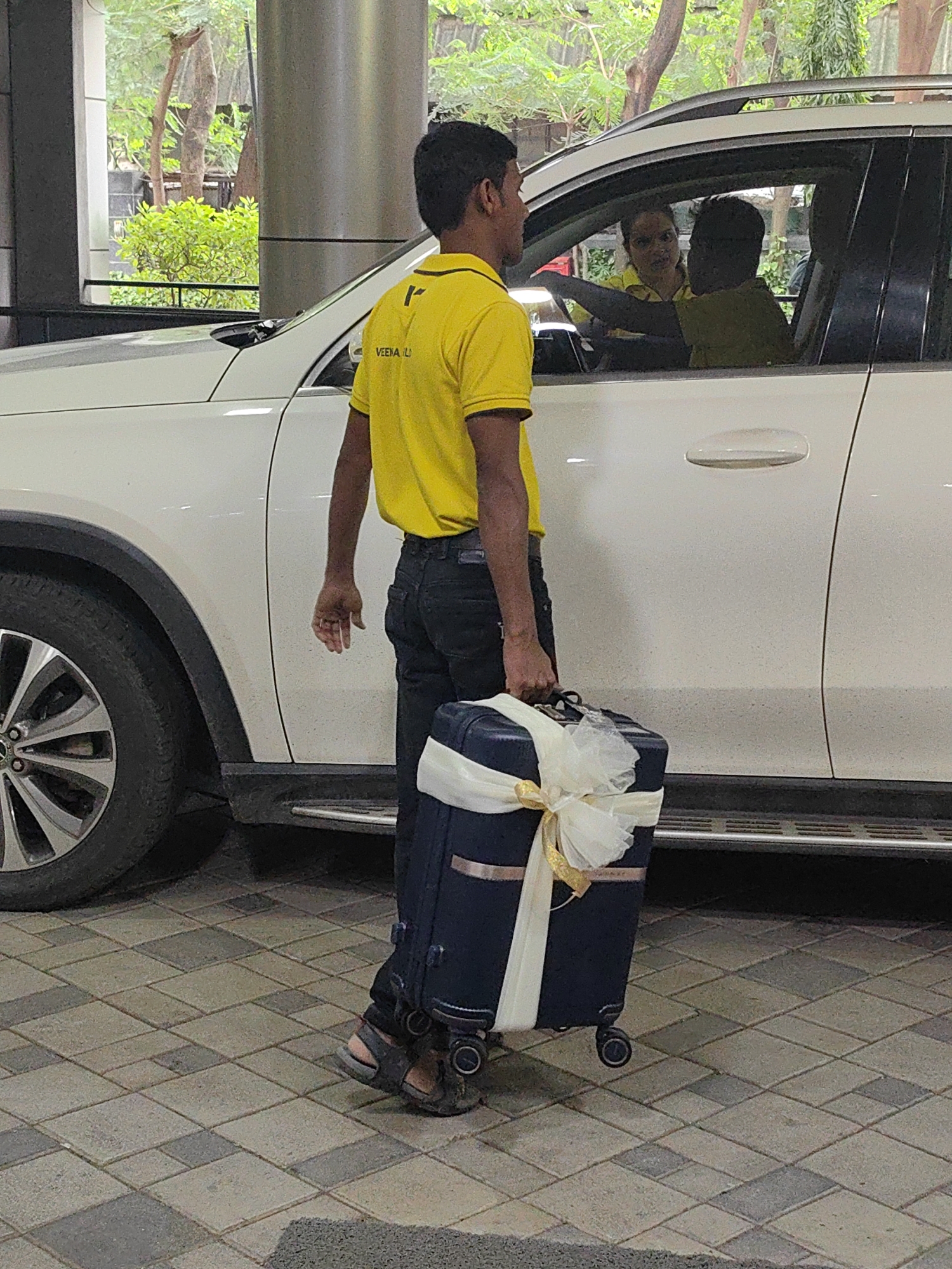 A person wearing a yellow t-shirt and black pants is walking next to a white SUV in Mumbai, India. The person is carrying a dark blue suitcase decorated with a cream-colored ribbon and bow. Inside the SUV, two people are visible; one is in the driver's seat and the other is in the passenger seat. The ground is paved with interlocking bricks. Trees and foliage are visible in the background.