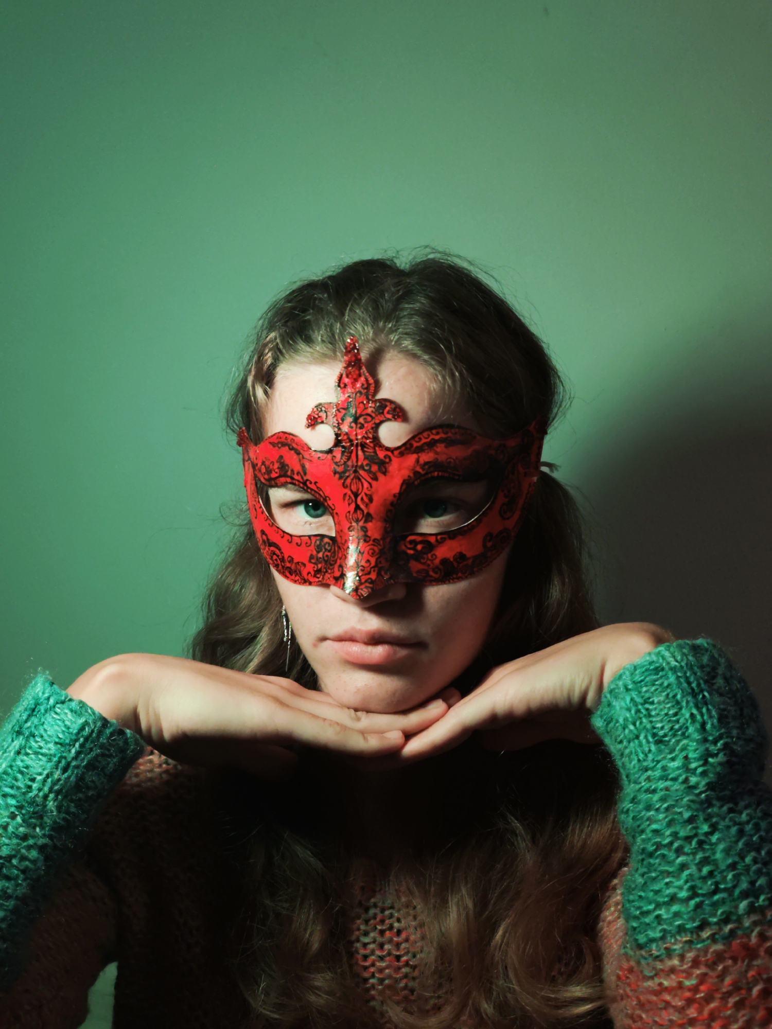 A person is seen from the chest up, wearing a red masquerade mask with black ornate patterns. The person has shoulder-length wavy hair and light skin. They are wearing a green and brown knitted sweater, with their hands placed under their chin in a resting pose. The background is a solid, somewhat muted green color. The scene does not appear to be in Budva, Montenegro.