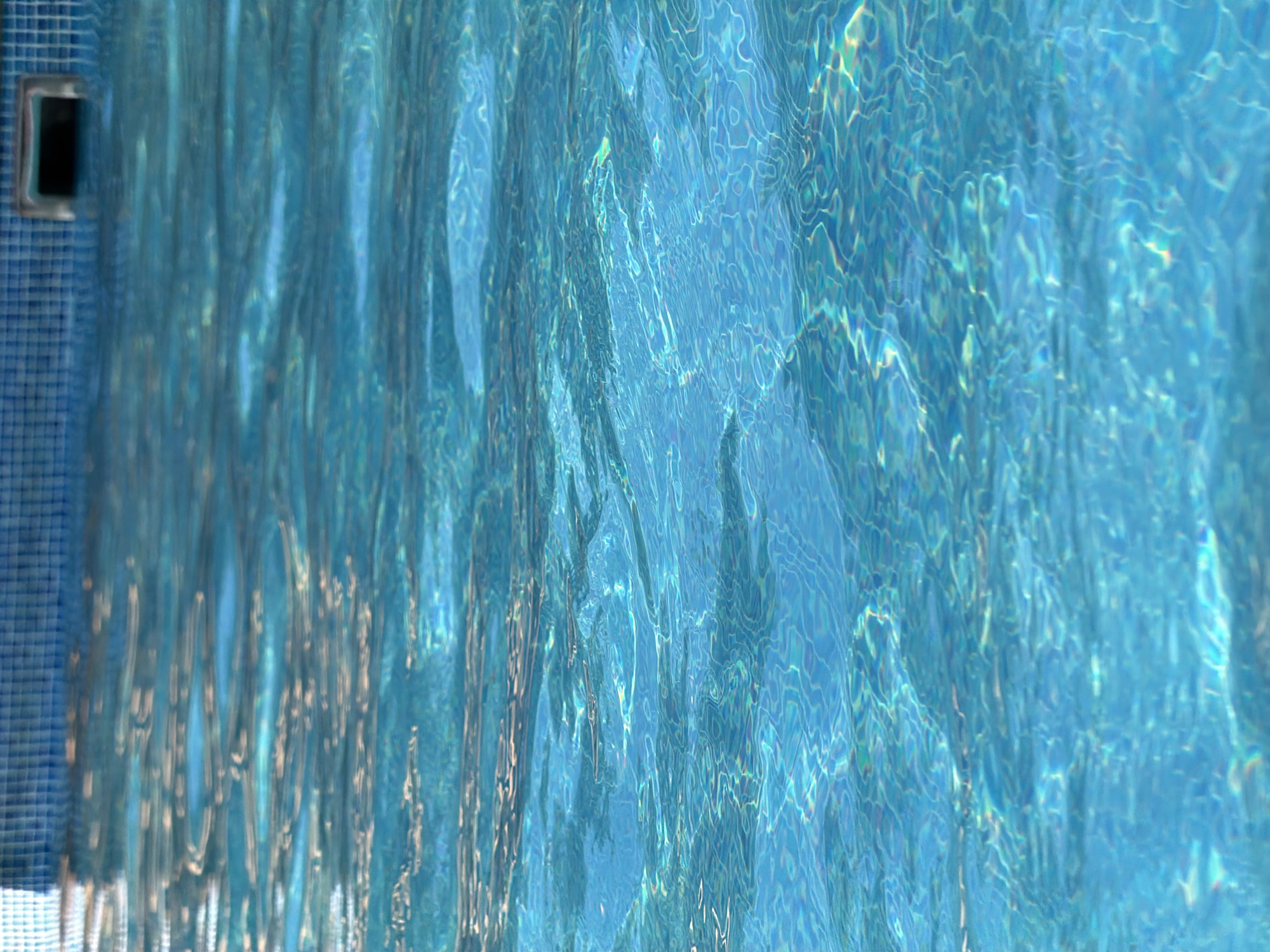 The image is a close-up, abstract view of the surface of a swimming pool. The water is a vibrant blue with shimmering reflections and patterns caused by light interacting with the ripples on the surface. The texture of the water dominates the frame, creating a dynamic and fluid visual effect.
On the left side of the image, a small portion of the pool's tiled edge is visible, featuring a grid of small, dark blue tiles. Integrated into this tiled area is a rectangular grate or drain, framed by a metallic border. This is the only distinct object in the scene, anchoring the otherwise abstract watery surface.
The lighting suggests it could be daytime, as indicated by the bright reflections and the clarity of the water. There are no people or animals visible, nor any specific cues about the weather beyond the tranquil state of the water. The scene evokes a sense of calm and coolness.
The image does not contain any visible text. The overall impression is one of the dynamic interplay of light and water, with a hint of the man-made structure of the pool providing context.