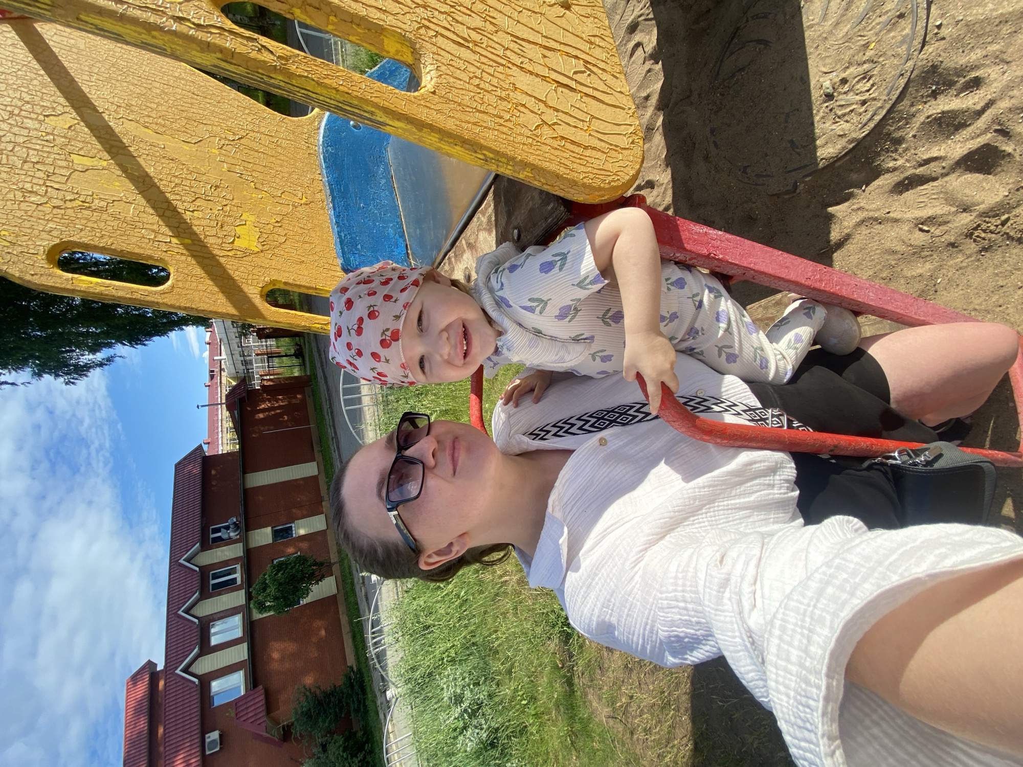 A woman and a young child are on a playground, possibly in Chaikovsky, Russia, as suggested by the background which includes a red-roofed building with cream-colored accents. The woman, wearing sunglasses and a white, textured shirt, is holding the child. The child, dressed in a light-colored outfit with a floral print and a red bandana with a cherry pattern, is smiling and sitting on a red play structure. The playground surface is sand, and a portion of a yellow and blue play equipment is visible. The sun is shining, casting shadows.