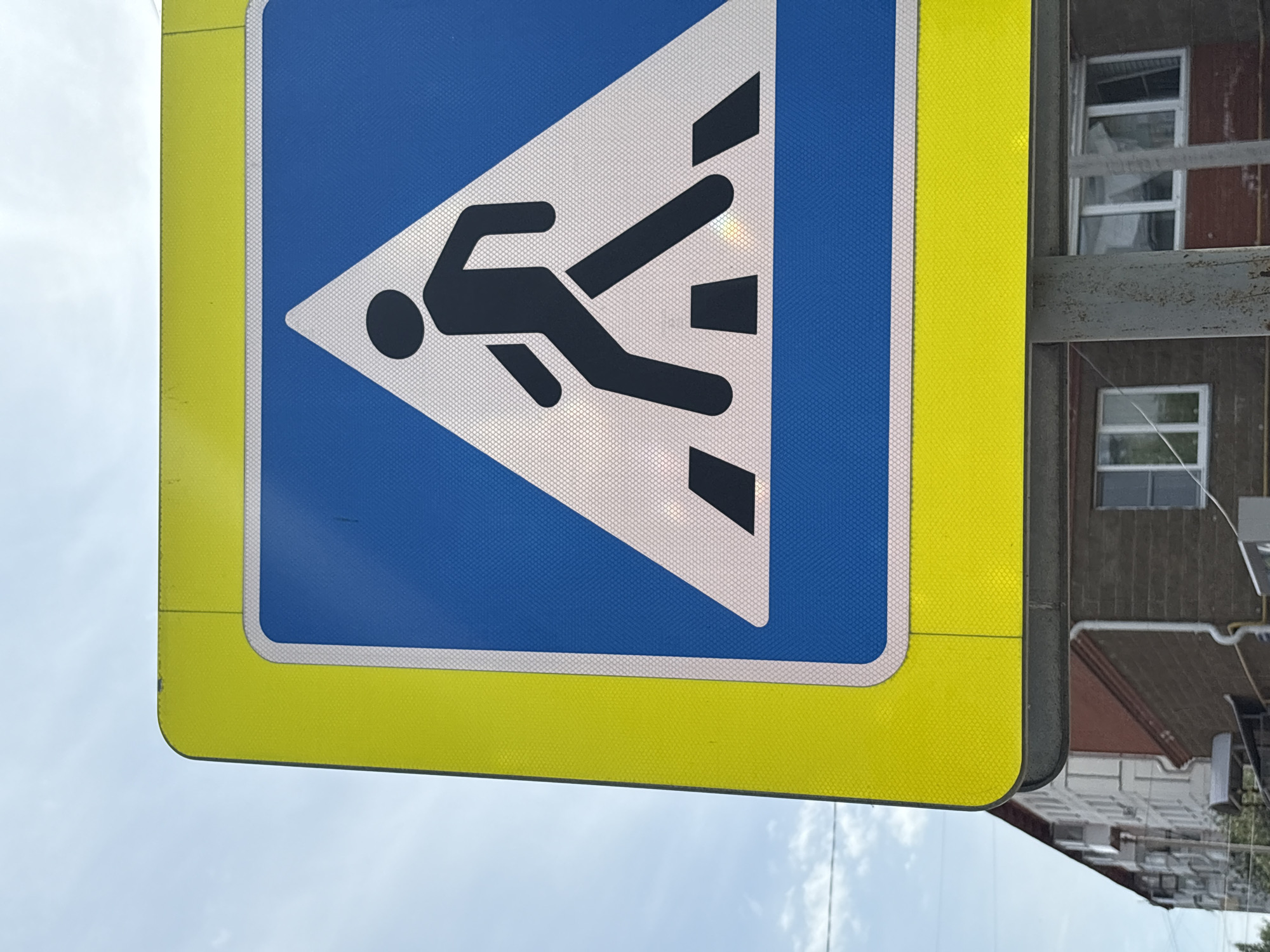 A road sign with a yellow border and blue interior displays a white triangle containing a running stick figure with appendages trailing behind, possibly indicating motion. The sign is attached to a gray metal pole. In the background, a building with white-framed windows is partially visible. The sky is visible in the bottom left corner. The specific location is Chaikovsky, Russia.