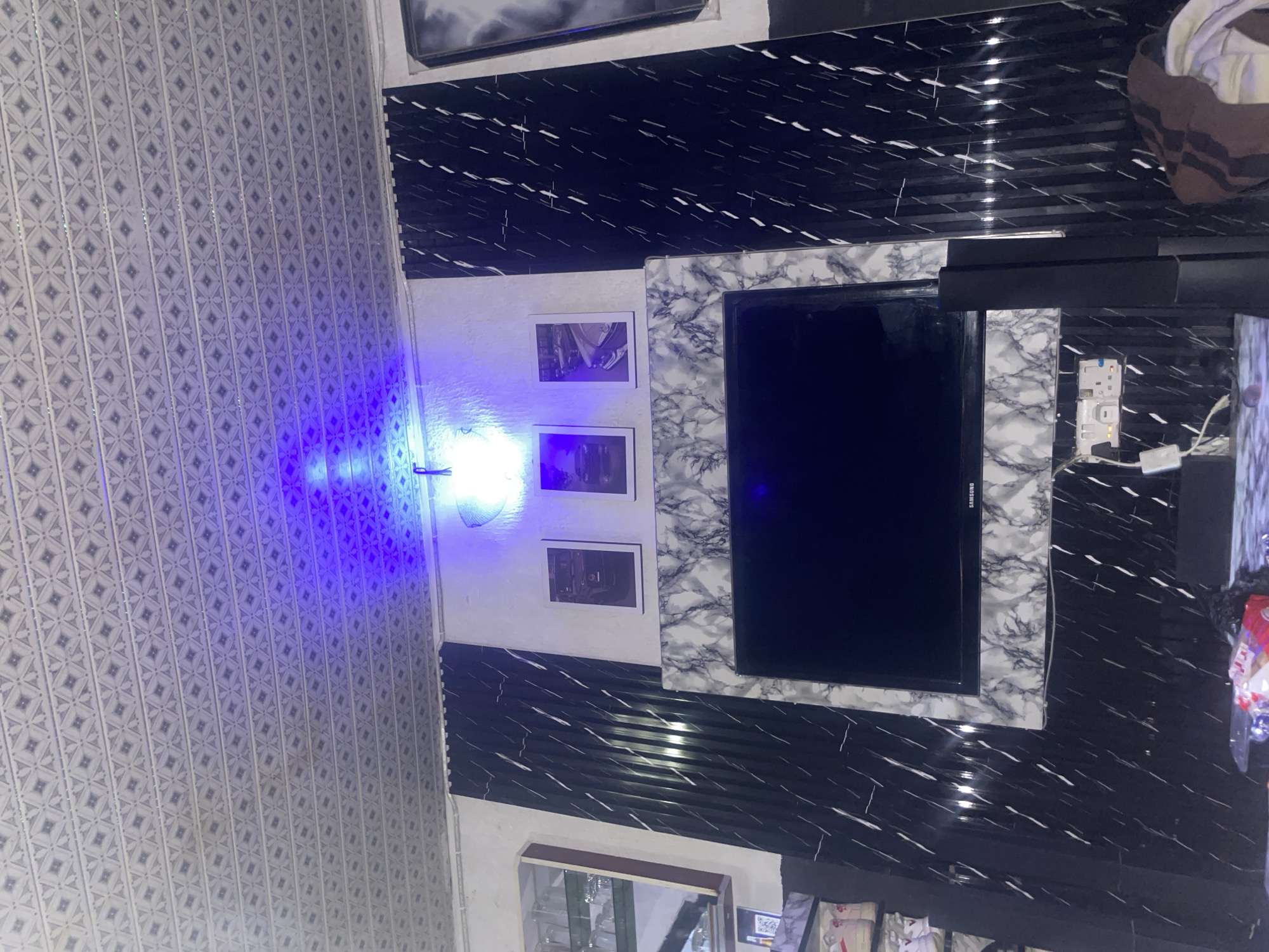 The image shows an indoor scene, likely a living room, with a television mounted on a wall. The wall features a decorative marble-patterned surround for the television, with black, vertically ribbed panels on either side. Above the television, there are three framed pictures on the wall, each depicting a car. A blue light emanates from the ceiling, casting a glow on the wall and the patterned ceiling panels, which appear to be made of a metallic material with a repeating geometric design. To the left of the television, a glass-fronted cabinet is partially visible, containing glassware. Below the television, a power strip with multiple outlets is mounted on the wall, with wires connected to it. A portion of a speaker system is visible to the right of the television. In the foreground, a table with some items on it is visible, including what appears to be a packaged item of food. The overall impression is of a decorated interior space, possibly intended for entertainment.