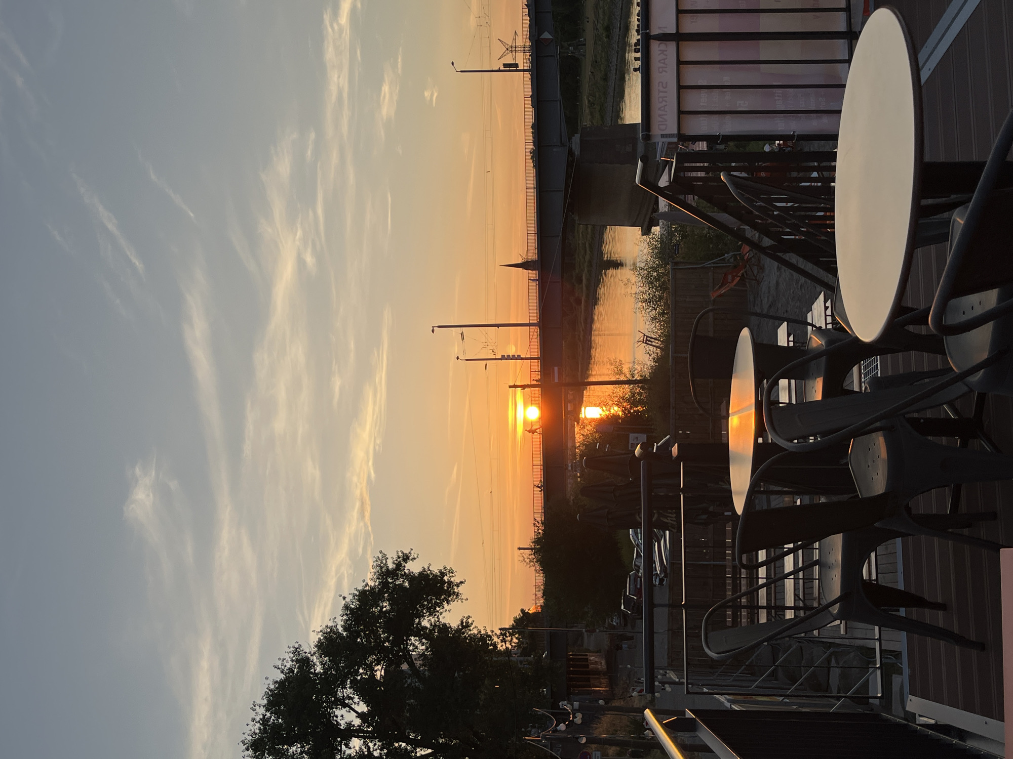 A low-angle, vertical shot captures a sunset over a river and a bridge, with tables and chairs in the foreground. The sun, a bright orange orb, is partially obscured by the horizon and reflected in the water. A bridge spans the river, with lampposts and overhead wires visible. On the left, a large tree with dark foliage is silhouetted against the sky. The foreground features outdoor seating with round tables and dark metal chairs, set on a wooden deck. The overall ambiance is serene and tranquil.