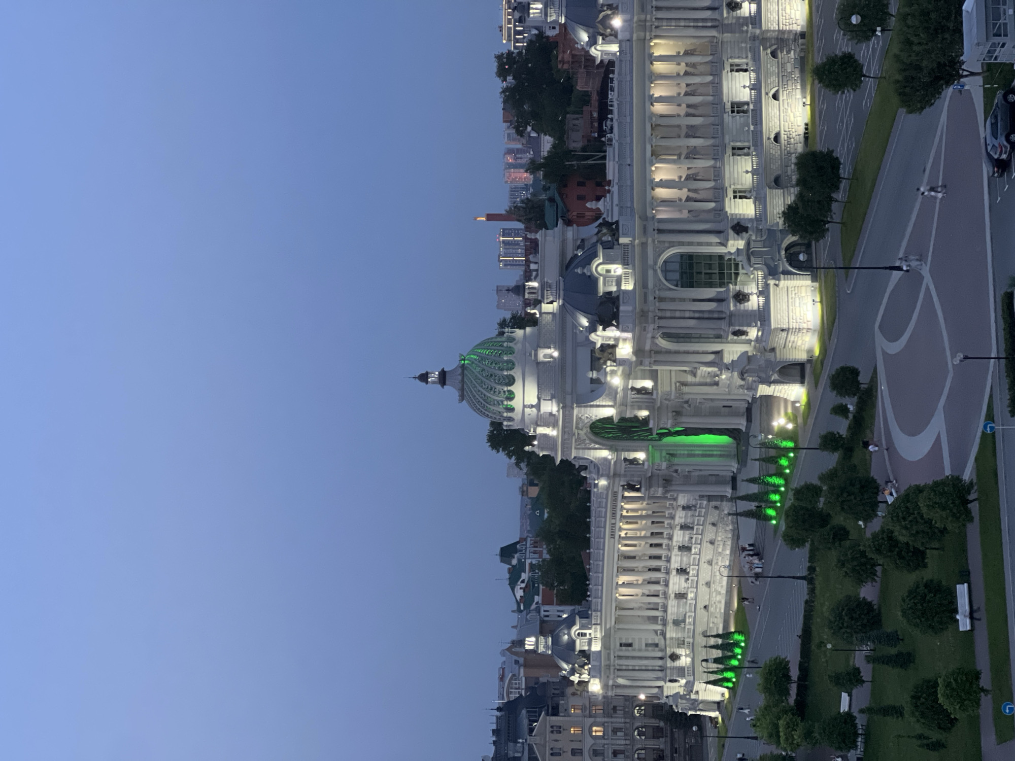 The image depicts the Palace of Farmers in Kazan, Russia at twilight. The palace, brightly lit, features a prominent green dome and ornate white facade. Adjacent to the palace is a paved area with trees, benches, and streetlights also illuminated. In the background, a skyline of buildings is visible, indicating an urban setting. The sky is a clear, deep blue, suggesting the time of day.