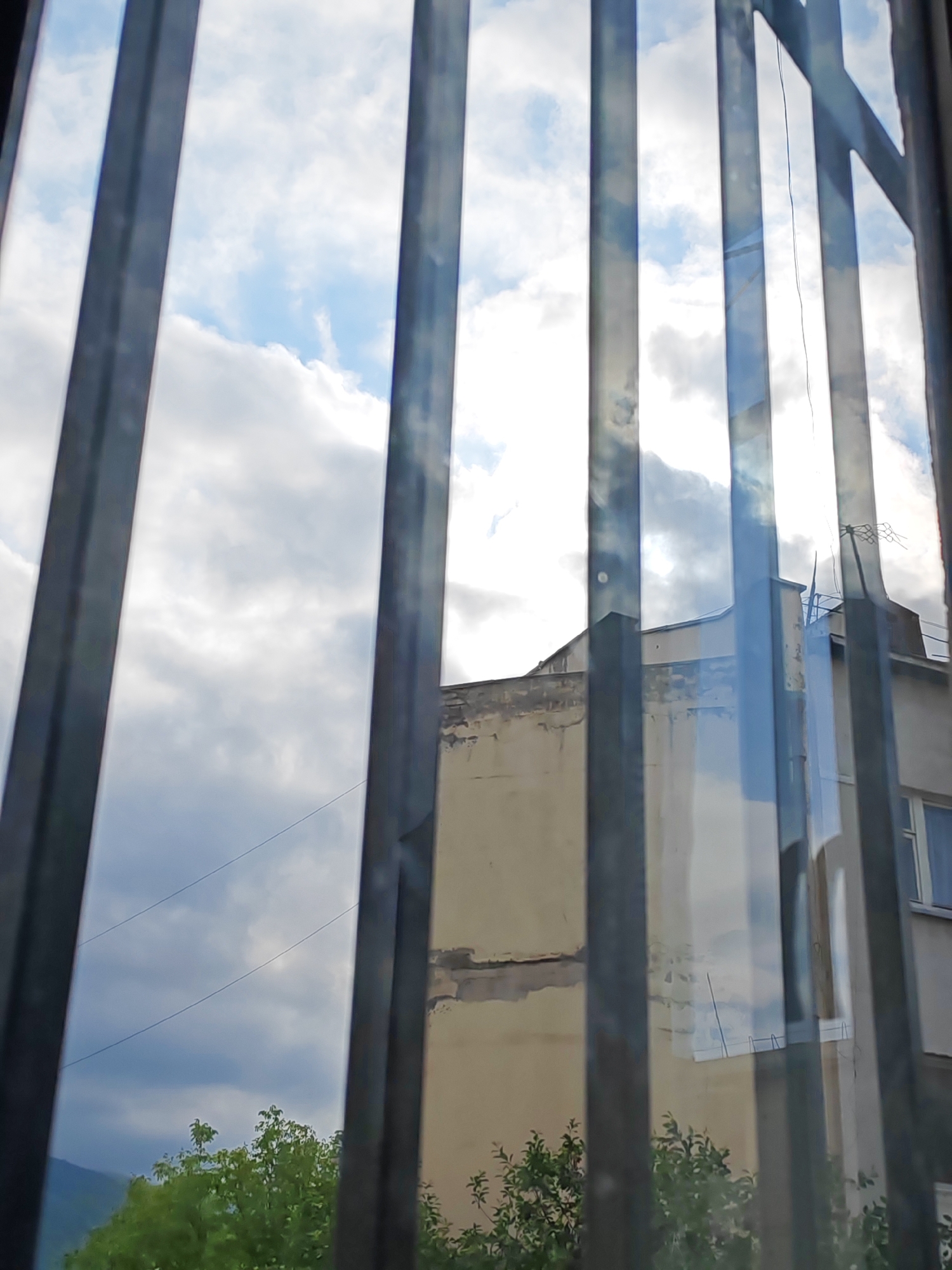 This image, taken in Vanadzor, Armenia, shows a view through metal bars, likely from a window or a protective barrier. The perspective is looking outwards towards a cloudy sky with patches of blue. In the middle ground, the facade of a building is visible, predominantly a light beige color with some weathered areas and visible structural damage. The building appears to be several stories high and has windows. In the foreground, the dark, vertical metal bars dominate the frame, creating a sense of confinement. To the left of the building, a portion of green trees is visible. In the far background, faint outlines of hills or mountains can be discerned. The lighting suggests it is daytime, with the clouds creating varied light and shadow on the scene. There are no people or animals depicted in the image.