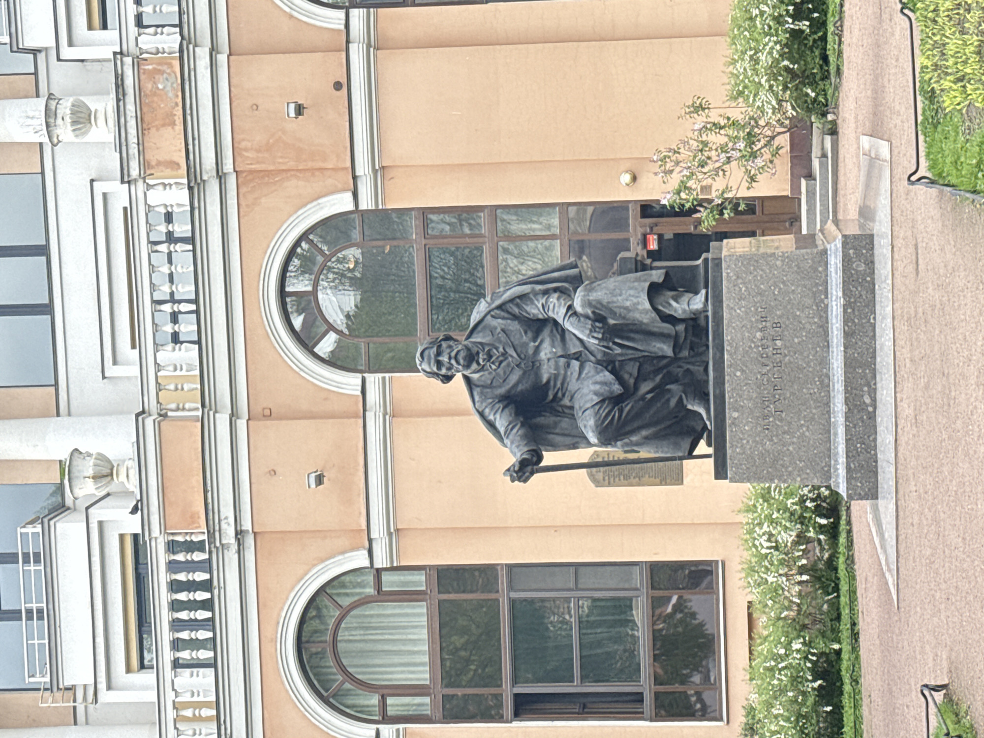 The image captures the monument to Ivan Turgenev in Saint Petersburg, Russia. The bronze statue of the seated writer is set against the backdrop of a light-colored building with arched windows and classical architectural details. The statue itself sits on a stone pedestal inscribed with the name "Иван Сергеевич Тургенев" (Ivan Sergeyevich Turgenev). The immediate surrounding area includes greenery, with bushes and a paved pathway visible. The architectural style and landscaping suggest a formal, urban park or square setting.