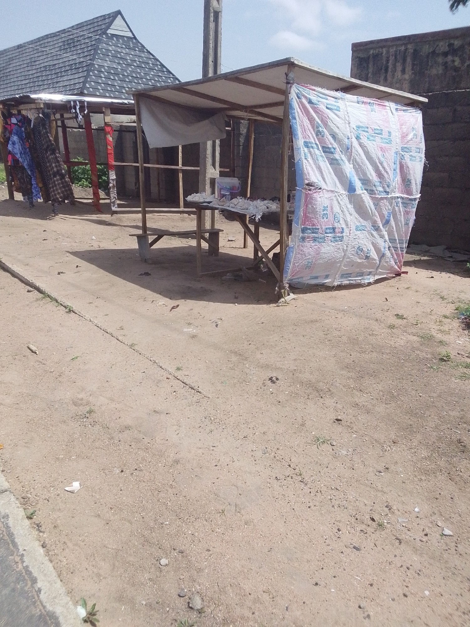 The image depicts an outdoor scene in Jalingo, Nigeria, featuring two makeshift market stalls. The stall on the left has a dark, shingled, peaked roof and displays various colorful fabrics hanging from a rail. The stall on the right is covered with a large, white tarp printed with red and blue lettering, likely from packaging. Beneath the tarp, wooden supports form a simple structure, and a table is visible with items on it, possibly goods for sale, appearing as small, light-colored objects. The ground is unpaved, composed of dirt and small stones, with a raised concrete edge running along the bottom left of the frame. A concrete pole stands to the right of the right stall. The background shows a rough brick wall and a hazy sky. There are no people actively engaged in the scene, but shadows suggest the presence of sunlight.