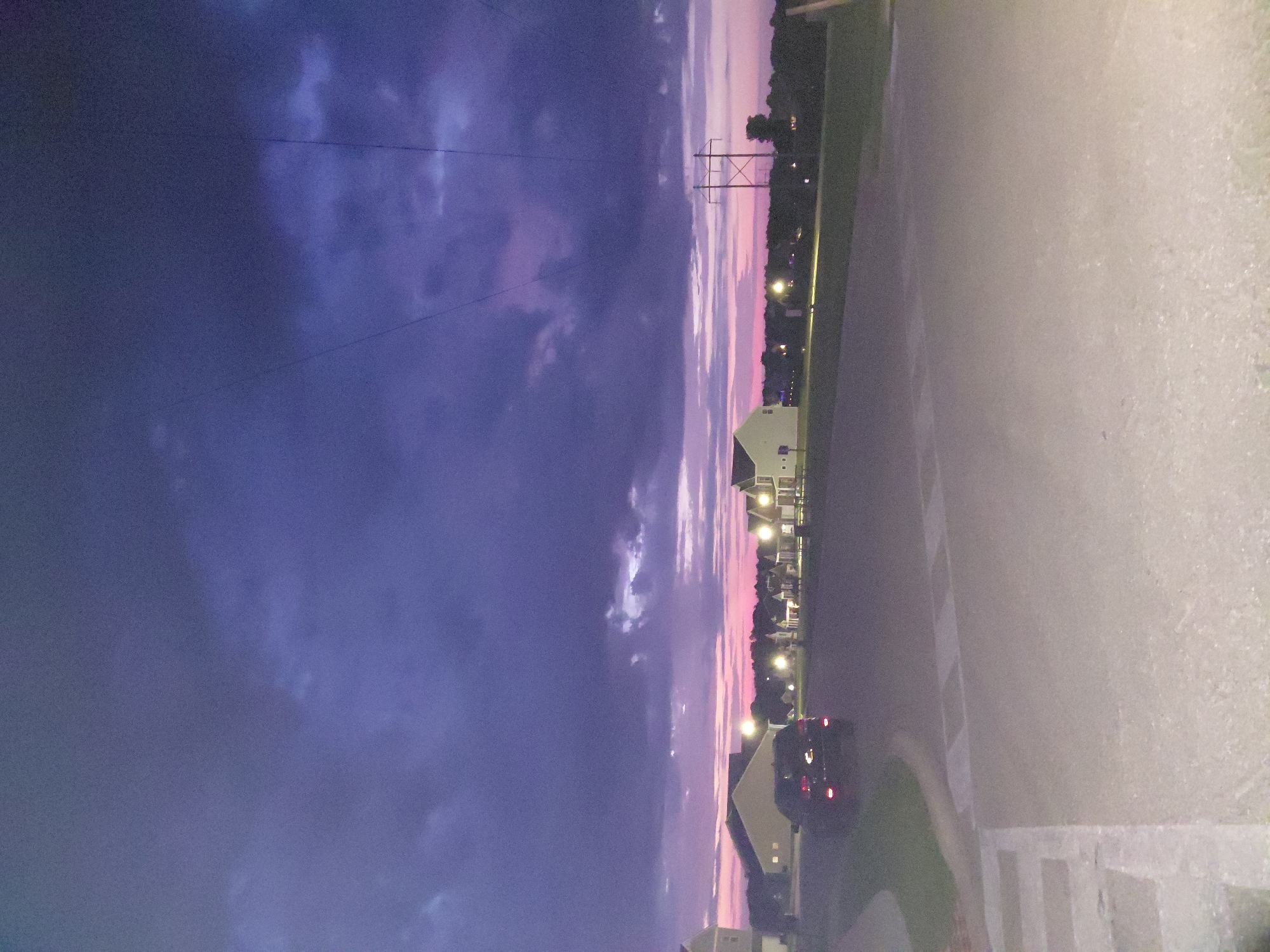 The image shows a suburban street scene at dusk in the city of Richmond, United States of America. The sky is a dramatic mix of deep purple and pink clouds, illuminated by the fading light of the sun. In the foreground, a paved road with white markings leads into the scene. A dark-colored SUV is parked on the left side of the road. In the mid-ground, there are several residential houses with illuminated windows, typical of a suburban neighborhood. Some streetlights are also visible, casting a warm glow. Further in the background, a line of trees separates the residential area from what appears to be a more open landscape. Power lines and a utility pole are visible against the twilight sky, indicating the presence of infrastructure. The overall impression is one of a quiet evening in a residential area.