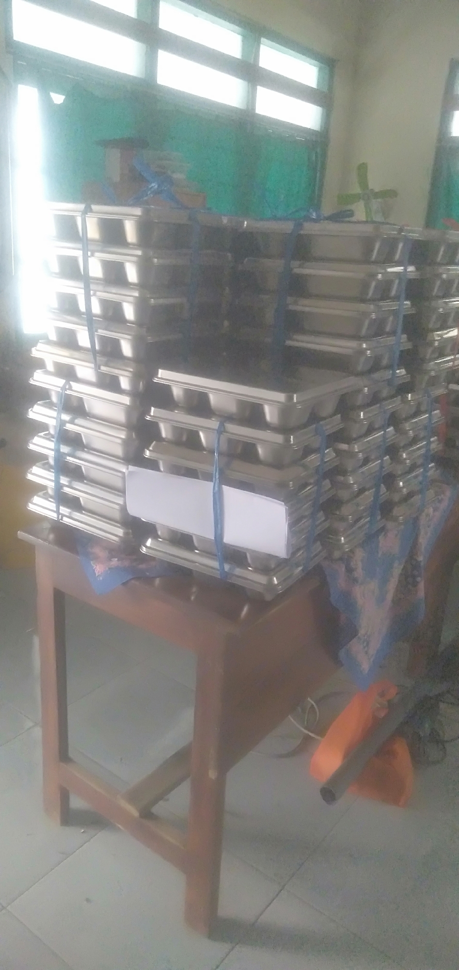 This image shows stacks of metal trays on a wooden table. The trays are rectangular with rounded corners and appear to be stacked in two columns. Blue straps are used to secure the stacks. A white paper or label is visible on one of the stacks. The table is made of wood with a simple four-legged design. In the background, there are windows with green curtains and a light-colored wall. There are also some electrical cords and an orange plastic bag on the floor. The scene appears to be indoors, possibly in a school or communal kitchen setting. The location is identified as Plemahan, Indonesia.