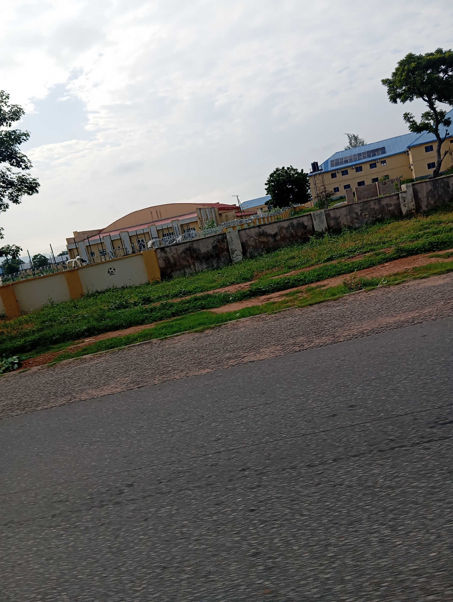 The image depicts a street view in Jalingo, Nigeria, featuring a section of a building with a yellow facade and a blue roof. A concrete fence, partially painted yellow and adorned with barbed wire, runs along the front of the property. Lush green vegetation covers the area between the fence and the street. The sky above is overcast with scattered clouds. The foreground is dominated by a textured asphalt road.