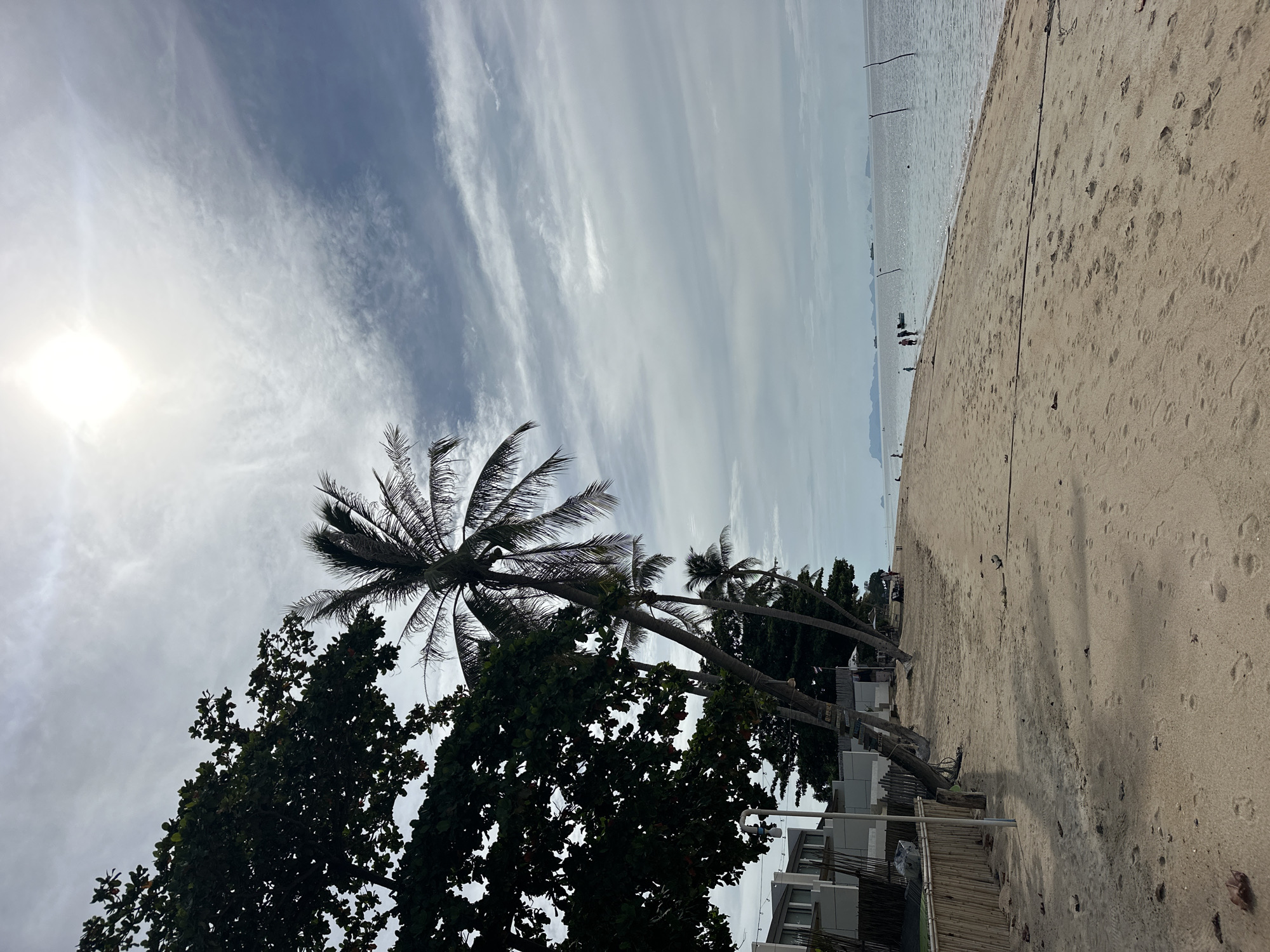 The image captures a serene beach scene in the Ko Samui District of Thailand. The foreground is dominated by a sandy beach with scattered footprints, leading to the gentle lapping waves of the ocean. Palm trees, with their fronds reaching towards the sky, line the edge of the beach, casting dappled shadows on the sand. Behind the palm trees, a row of modern white buildings with dark-framed windows suggests a resort or residential area. A bamboo fence runs along the property line. In the distance, several small boats are visible on the calm water, and a hazy outline of islands can be seen on the horizon. A few individuals are present on the beach, some walking near the water's edge, adding a sense of scale and life to the tranquil landscape. The sky is partly cloudy, with the sun peeking through, creating a bright, diffused light that illuminates the scene.