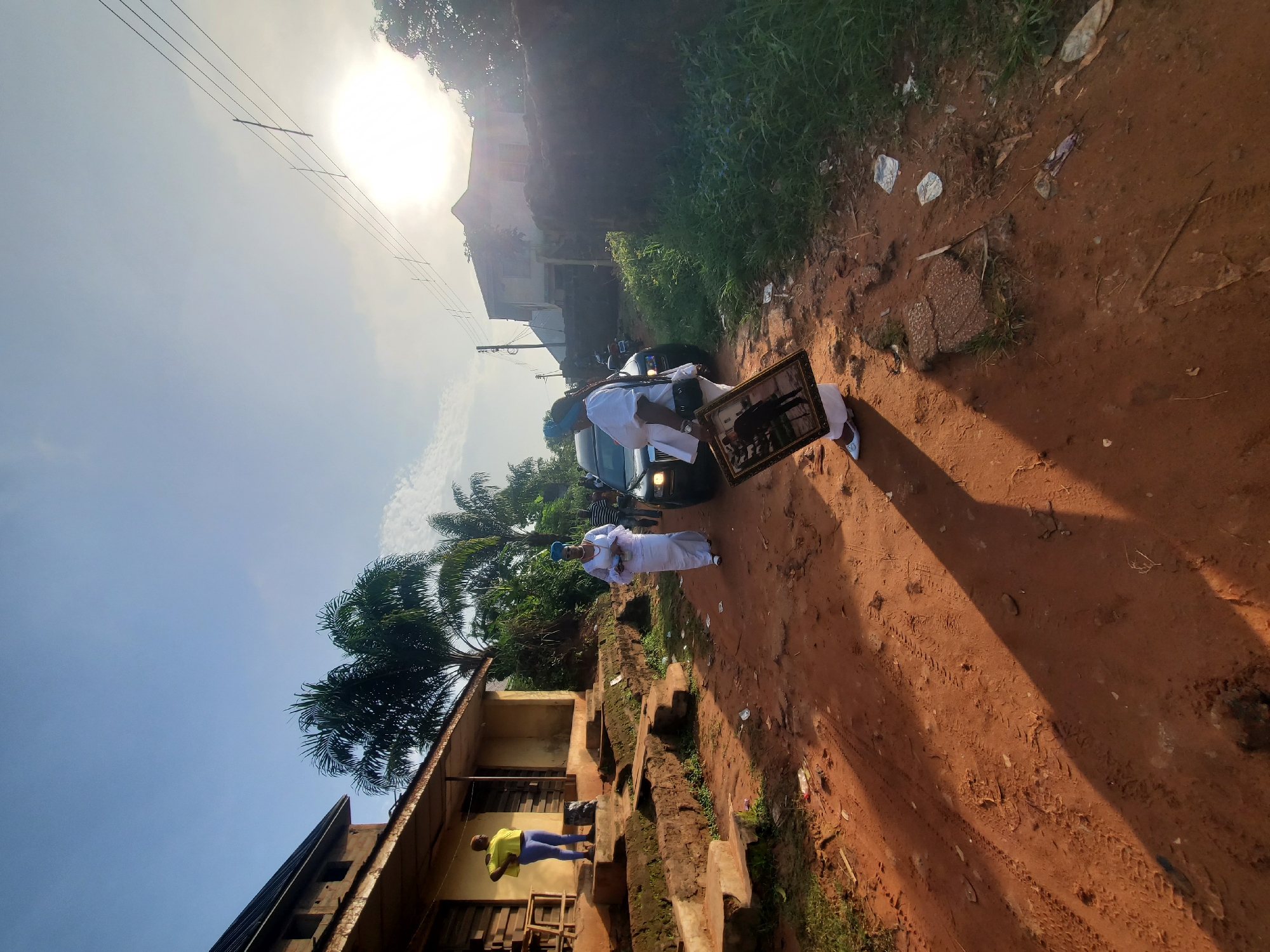 In Obosi, Nigeria, a group of people are gathered on a red dirt road. At least two individuals, wearing white ceremonial attire, are visible. One is holding a framed photograph. A dark car is nearby. The surrounding environment includes vegetation, a multi-story building with a person standing on its steps, and power lines above. The sun is high in the sky. The setting appears to be a rural or residential area within the city.