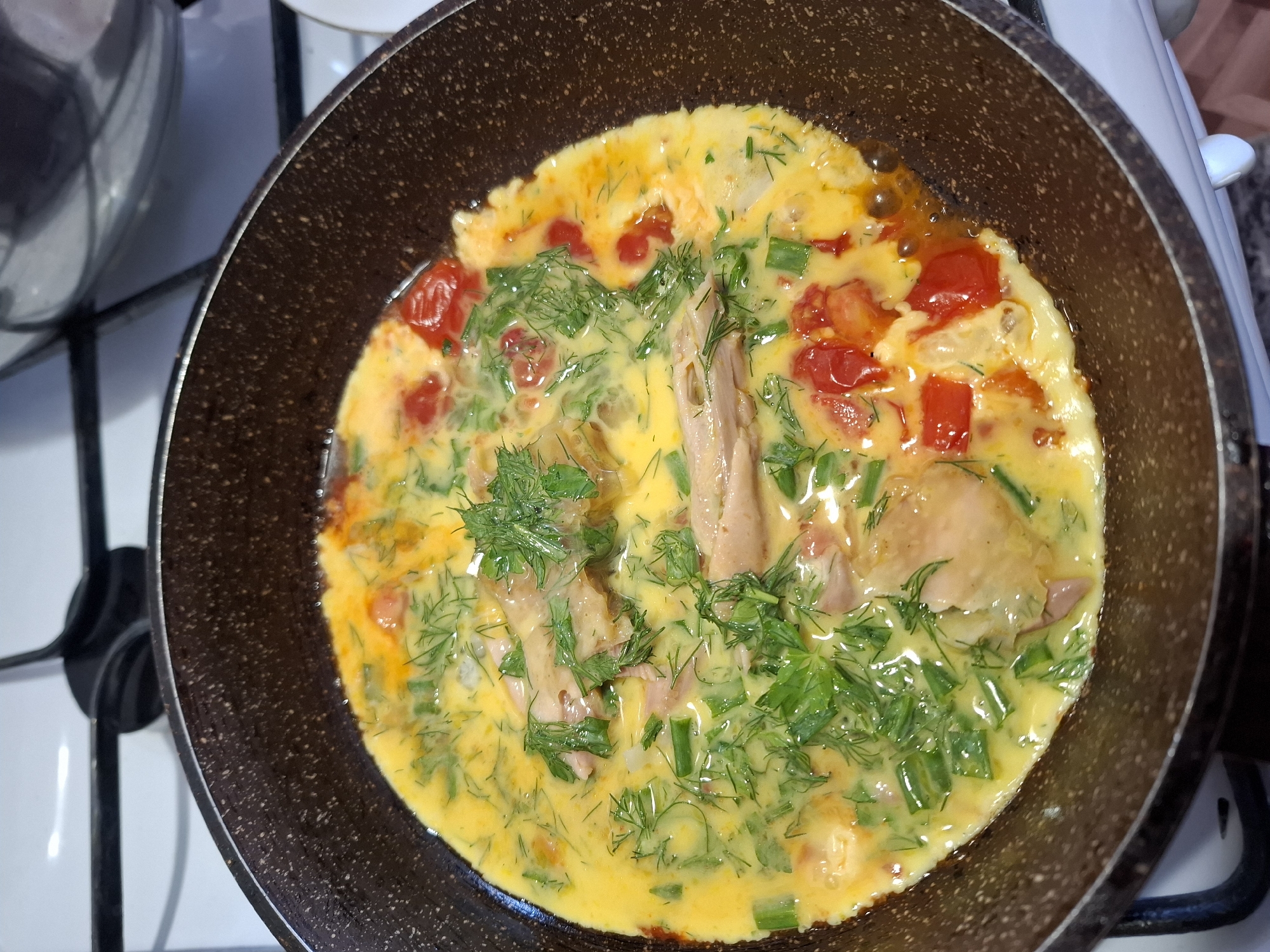 The image shows a close-up, overhead view of an omelet being cooked in a dark-brown non-stick frying pan. The omelet appears to be made with eggs, chopped cherry tomatoes, green onions, and dill. There are also pieces of what appear to be cooked chicken or fish within the omelet. The omelet is still in the process of cooking, with some liquid egg visible. The frying pan is on a stovetop, with the edge of a white stovetop visible in the background. The image was taken from a slightly elevated angle. The setting is a kitchen, likely in a residential home. The provided information indicates the location is Korenovsk, Russia.