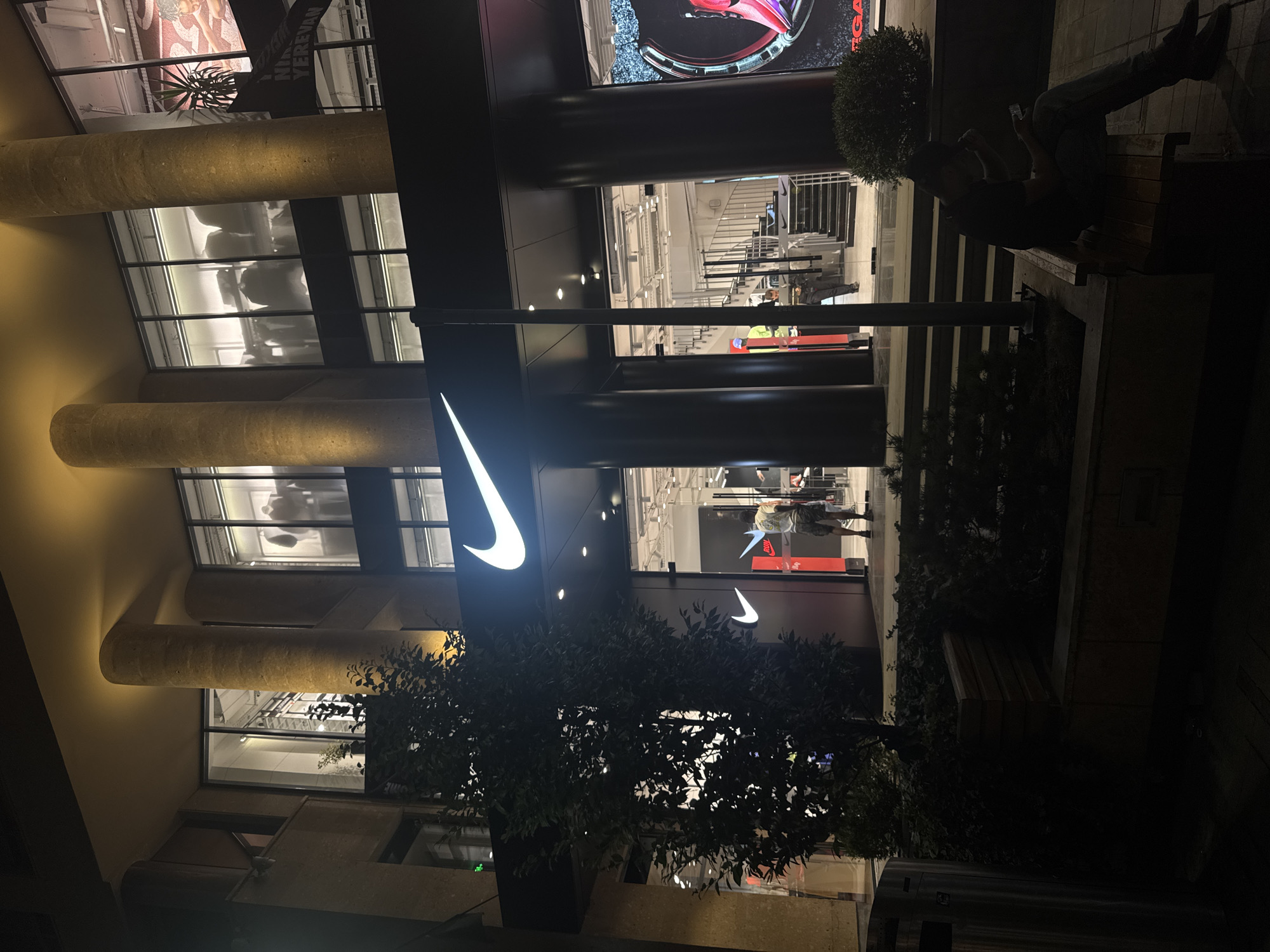 The image captures the exterior of a Nike store in Yerevan, Armenia, at night. The store features a prominent, illuminated Nike logo above its entrance, with large glass windows showcasing the interior. Several large, cylindrical columns support the building's facade. Inside, mannequins are visible displaying clothing, and a spiral staircase ascends within the store.
Outside the store, a person is seated on a wooden bench to the right, looking at their phone. Another individual, dressed in casual attire, stands near the entrance. A dark-leafed tree and some bushes are positioned in front of the store, partially obscuring the view. The lighting is a mix of the store's interior glow and streetlights, creating a somewhat dramatic ambiance. The ground appears to be paved.