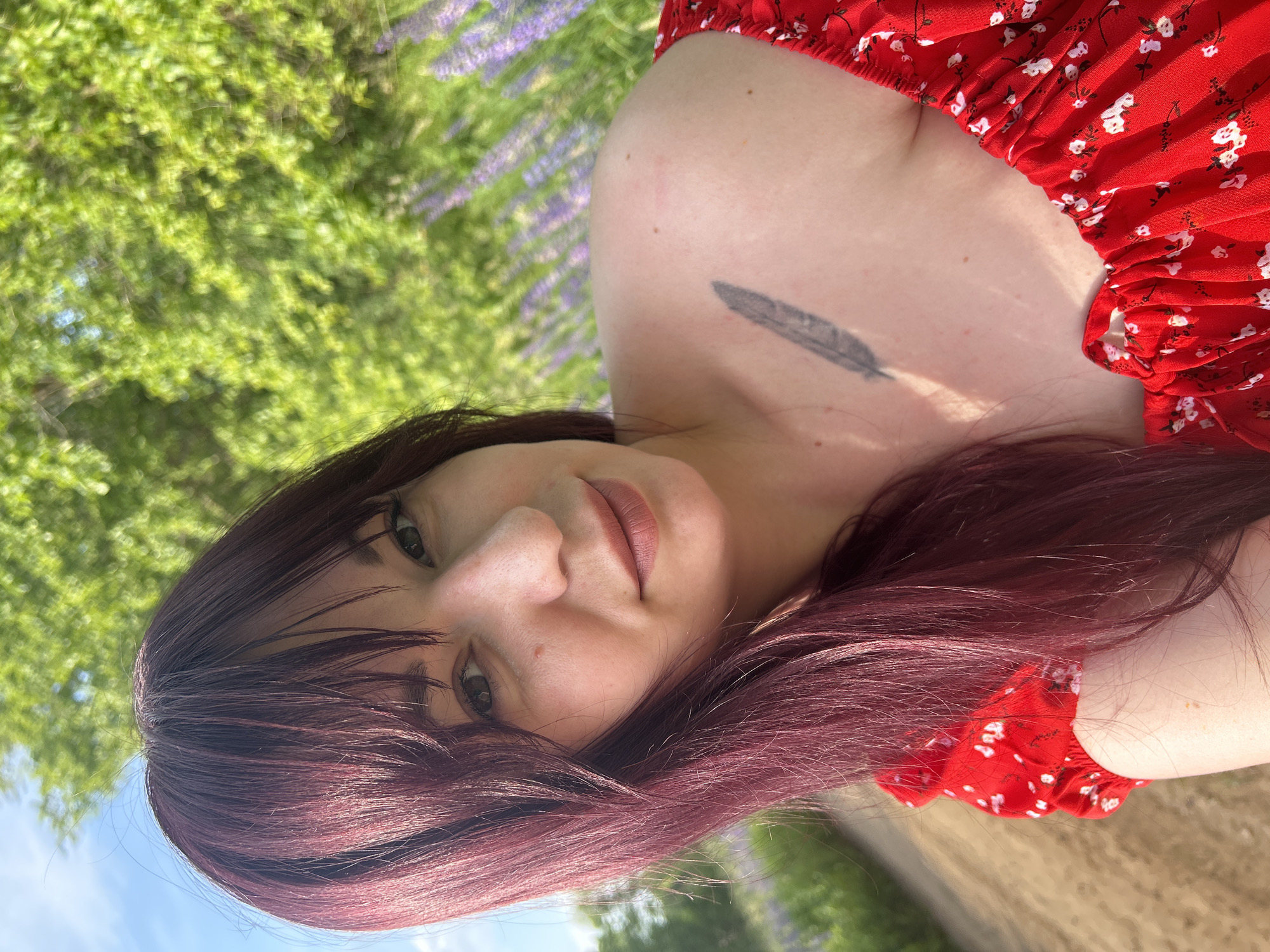 In a close-up, outdoor, selfie-style photograph, a woman with long, dark, reddish-brown hair and fair skin is the central subject. She is wearing a red off-the-shoulder dress with a white floral pattern. A black feather tattoo is visible on her left shoulder blade. The background consists of lush green foliage and a field of purple flowers, suggesting a rural or park-like setting. A dirt path is visible to the left of the woman. The lighting indicates it is daytime. The image appears to have been taken in the vicinity of Гаревая, Russia, given the mention of the location.
