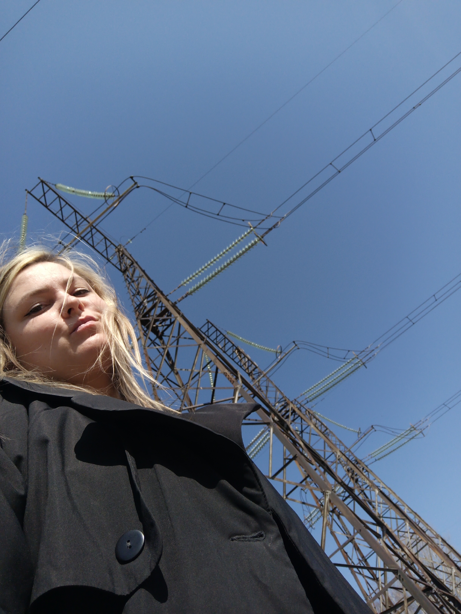 A low-angle, upward-facing shot features a young woman with blonde hair against a clear blue sky and a large electrical transmission tower. The woman is positioned in the lower-left portion of the frame, looking directly at the camera with a somewhat neutral, perhaps slightly pouting, expression. Her blonde hair is windswept, framing her face. She is wearing a dark, likely black, jacket or coat, with a prominent button visible on the front.
Behind her, a substantial metal lattice-work power pylon stands tall, its complex structure and multiple high-voltage transmission lines extending across the upper and right sides of the image. The lines are adorned with rows of light green or translucent ceramic disc insulators. The bright, clear blue sky indicates fair weather and daytime conditions, possibly mid-day due to the direct lighting.
The setting combines a personal portrait with the industrial landscape of power infrastructure, likely in or near Noviy, Russia, given the context. The photograph captures a moment of direct engagement from the subject against an imposing, yet functional, backdrop. There is no visible text in the image.