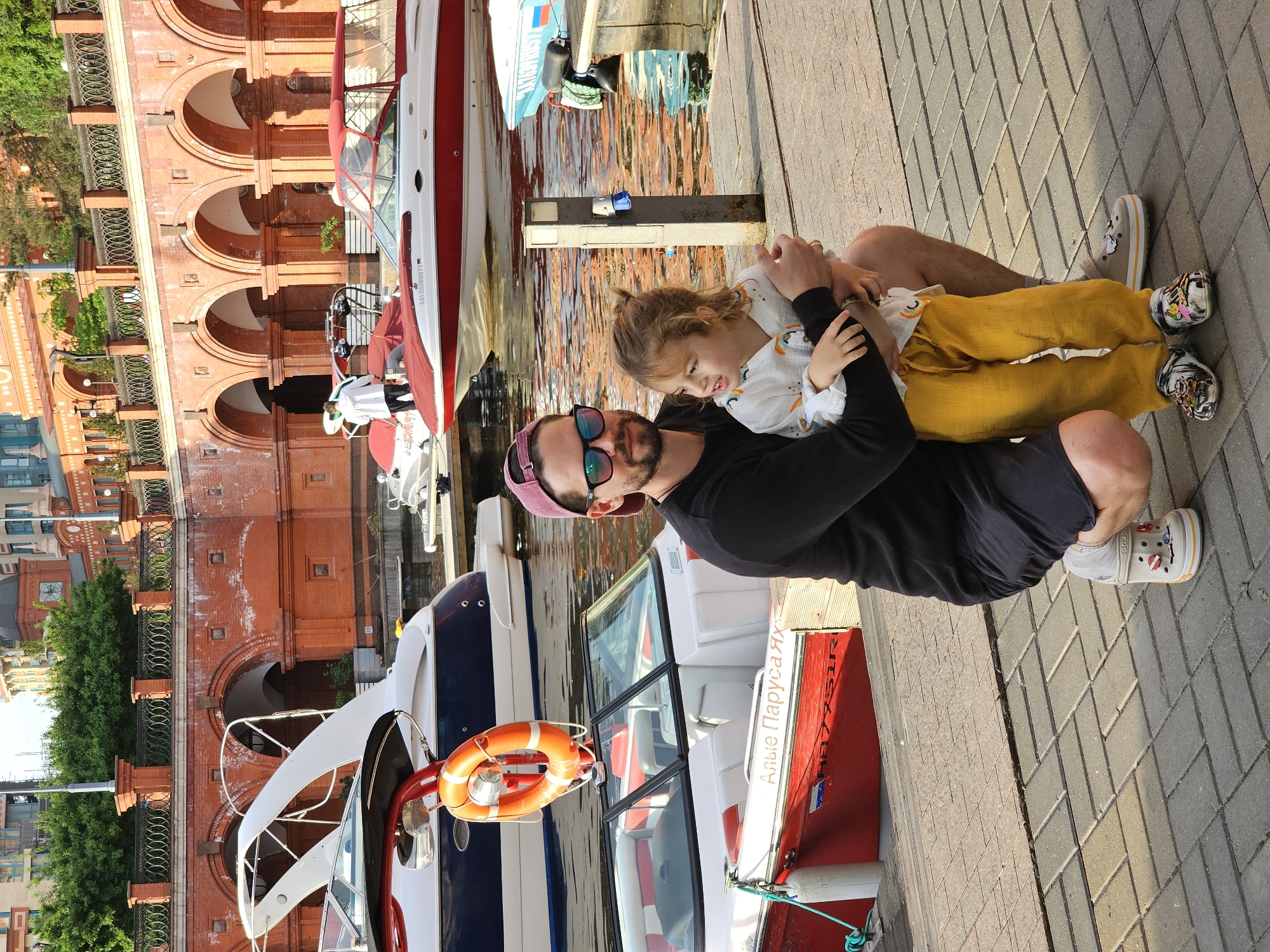 A man wearing a pink cap and sunglasses sits on a gray-brick pier with a child in his lap. The child wears yellow pants and a patterned white top. Several boats are docked beside the pier. A red brick building with archways is in the background. The boat closest to the foreground has an orange life preserver attached. The boat has some Russian writing on it, and the license plate visible is R977331R. The scene is in Khimki, Russia.