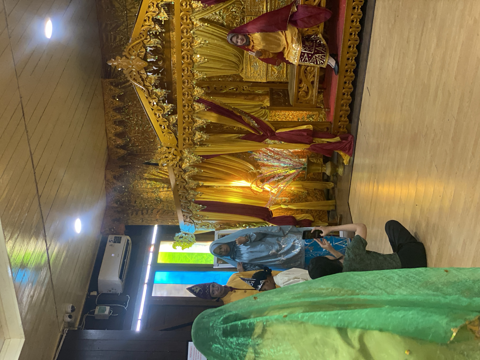 The image depicts an indoor scene, likely a cultural event or exhibit in Banjarmasin, Indonesia. In the foreground, a person is captured from behind, wearing a bright green headscarf and taking a photo with their phone. To their left, three individuals are dressed in traditional attire. A man wearing a yellow shirt and a purple patterned hat stands with a young child in a white shirt and a matching purple hat. Beside them, a young woman is in a light blue traditional outfit, complete with a headscarf.
The background features an elaborate, gold-colored throne adorned with intricate carvings and hanging ornaments, reminiscent of royal or ceremonial seating. Red and yellow drapes frame the throne, adding to the festive ambiance. Seated on the throne is a woman wearing a yellow dress and a dark red headscarf, with a patterned skirt. She is posing for the photograph, holding up two fingers in a peace sign.
The setting appears to be a well-lit room with a wooden ceiling and floor. An air conditioning unit is visible on a dark wall, along with security cameras. Stained glass windows in vibrant colors are also present, suggesting a unique architectural detail. The overall impression is one of a cultural celebration, possibly a traditional wedding or a museum display showcasing local heritage.