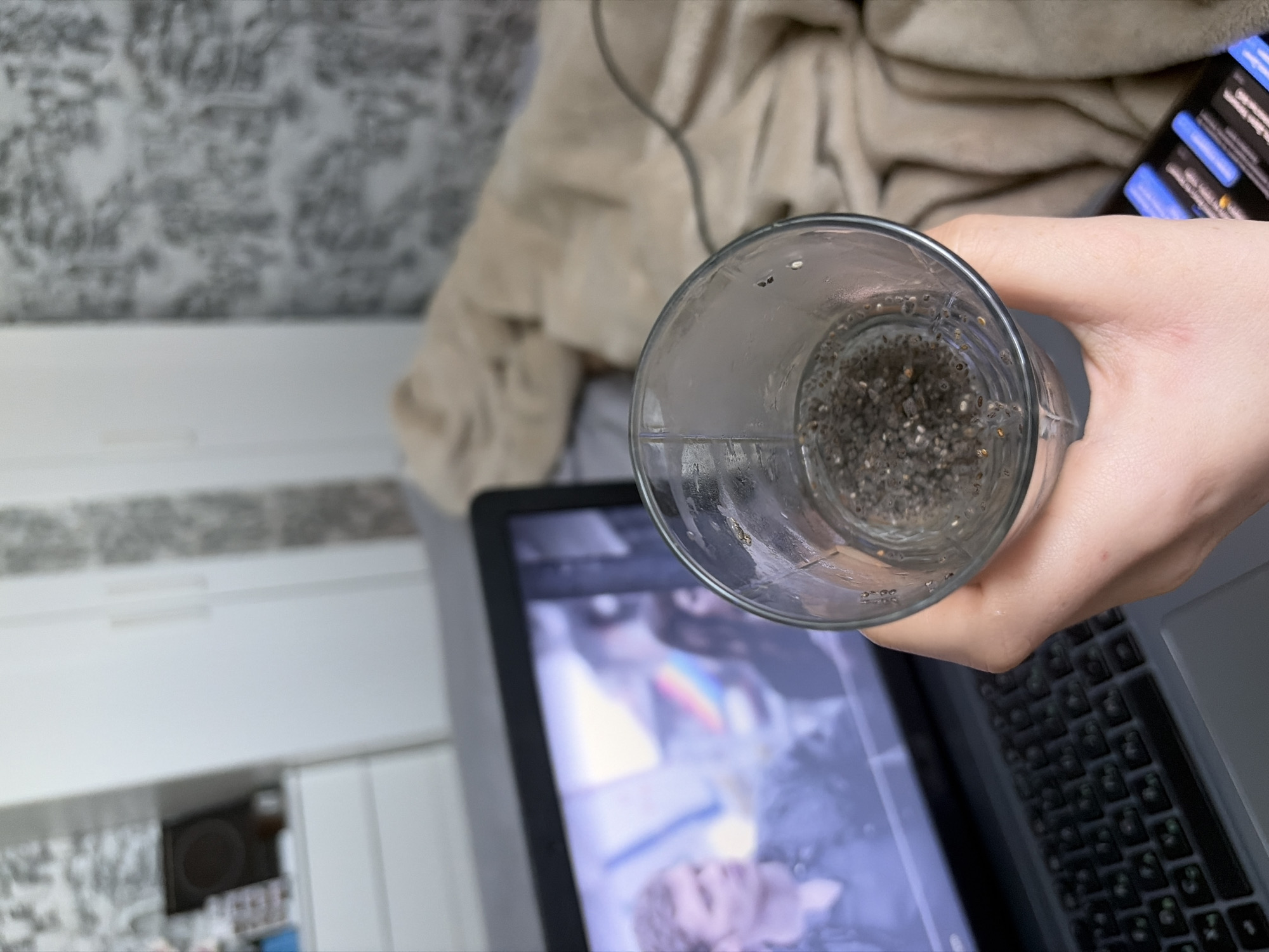 The scene depicts an individual holding a glass of liquid containing chia seeds while watching a video on a laptop, presumably in Новинки, Russia. In the foreground, a pale-skinned hand grips a clear glass filled with water and a concentrated layer of dark, small chia seeds at the bottom, with some seeds floating or adhering to the inner surface of the glass. The glass is positioned above a dark-colored laptop, which displays a video on its screen. The video features a male character with light brown, curly hair, wearing a dark shirt, prominently visible on the left side of the screen; this character strongly resembles actor Joseph Morgan. A rainbow-colored light streak is visible in the background of the video footage, alongside another partially visible individual to the right. The laptop's dark keyboard is partially visible beneath the hand and glass. To the right of the laptop, a plush, light-beige blanket or throw is draped across a surface. Below the blanket, a portion of a black smartphone screen displaying multiple blue-highlighted notifications or user interface elements is visible. In the background, to the left, white, minimalist furniture, possibly wardrobes or drawer units, is visible alongside a dark, circular object on a white surface. The walls throughout the room are covered with a patterned wallpaper in shades of grey and white.