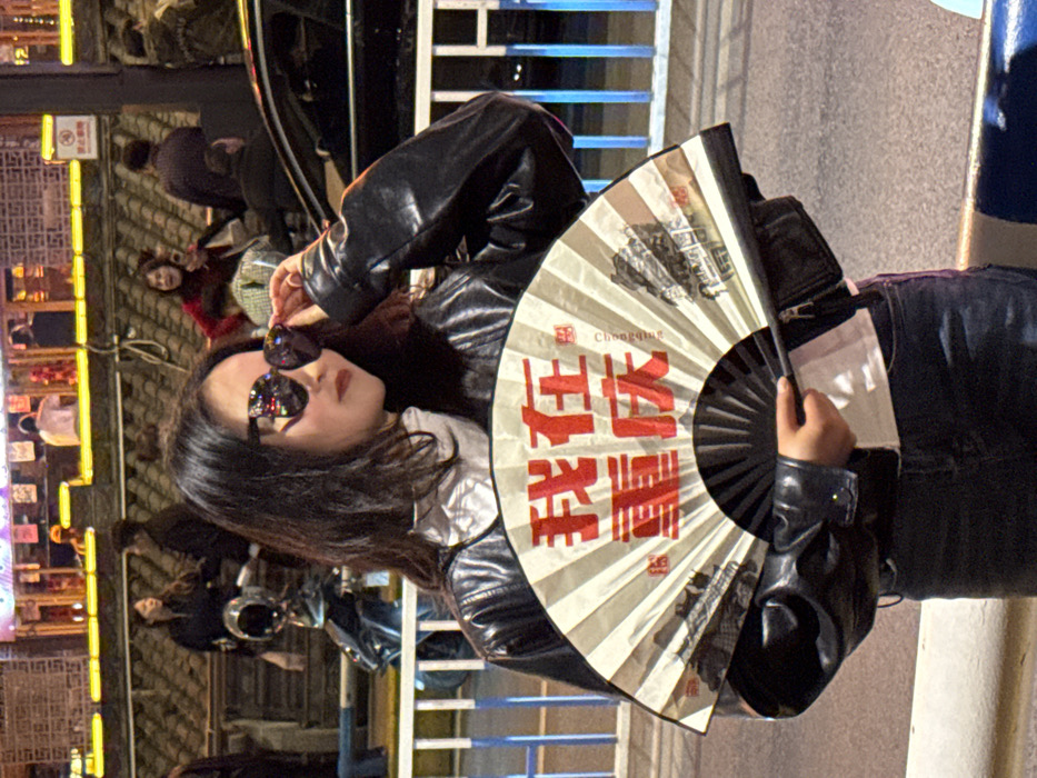 The scene is an urban street at night in Yuzhong District, Chongqing, China. A female individual, appearing to be of East Asian descent, stands facing slightly right of the viewer. She wears a black leather jacket over a white collared garment, dark trousers, and large, reflective sunglasses. Her left hand is positioned near the right temple of her sunglasses. She holds a partially opened traditional Chinese folding fan in her right hand. The fan's surface is beige with vertical stripes and features prominent red Chinese characters reading "我在重庆" (I am in Chongqing), alongside smaller English text "Chongqing" and two monochrome landscape illustrations. The immediate foreground features a blue and white striped barrier. The background is characterized by blurred figures, including individuals on foot and inside an elevated, brightly lit architectural structure featuring traditional Chinese design. A parked black scooter with a chrome helmet is visible to the left, and a dark-colored vehicle is partially visible to the right. A circular red and white warning sign is discernible in the upper right background.