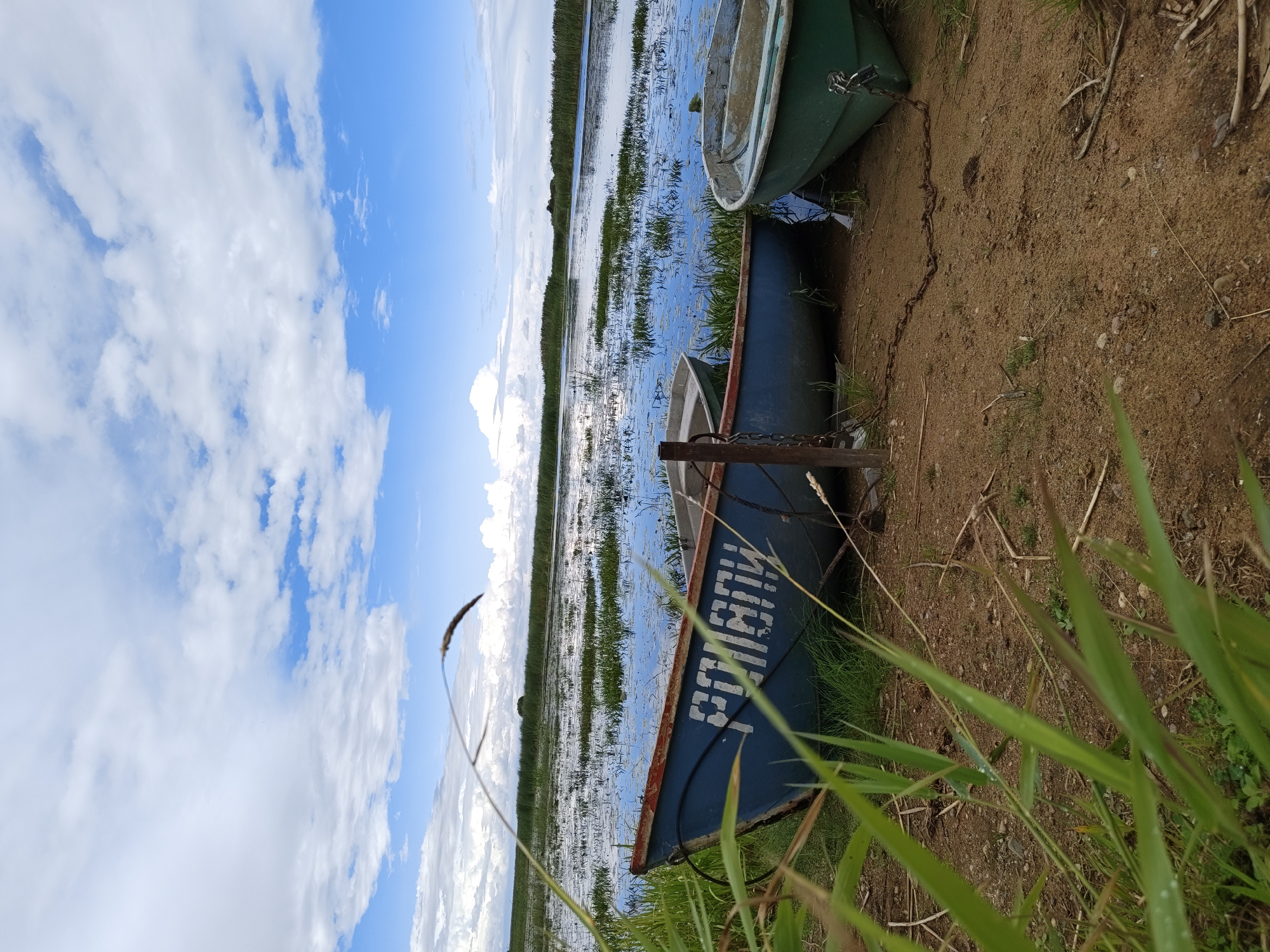 The image depicts a serene waterscape at the edge of Malaya Listovka, Russia, under a partly cloudy sky. Three boats are present on the sandy shore, two blue and one green, with the word "РОНАНИ" visible on the side of one of the blue boats. The boats are secured with chains to a metal post. The foreground is dominated by lush green grass and reeds, partially obscuring the view of the boats. The water is calm and reflects the sky, with patches of aquatic vegetation dotting its surface. In the background, a dense line of reeds stretches across the horizon, suggesting a marshy or lake environment. The overall scene conveys a sense of peacefulness and natural beauty.