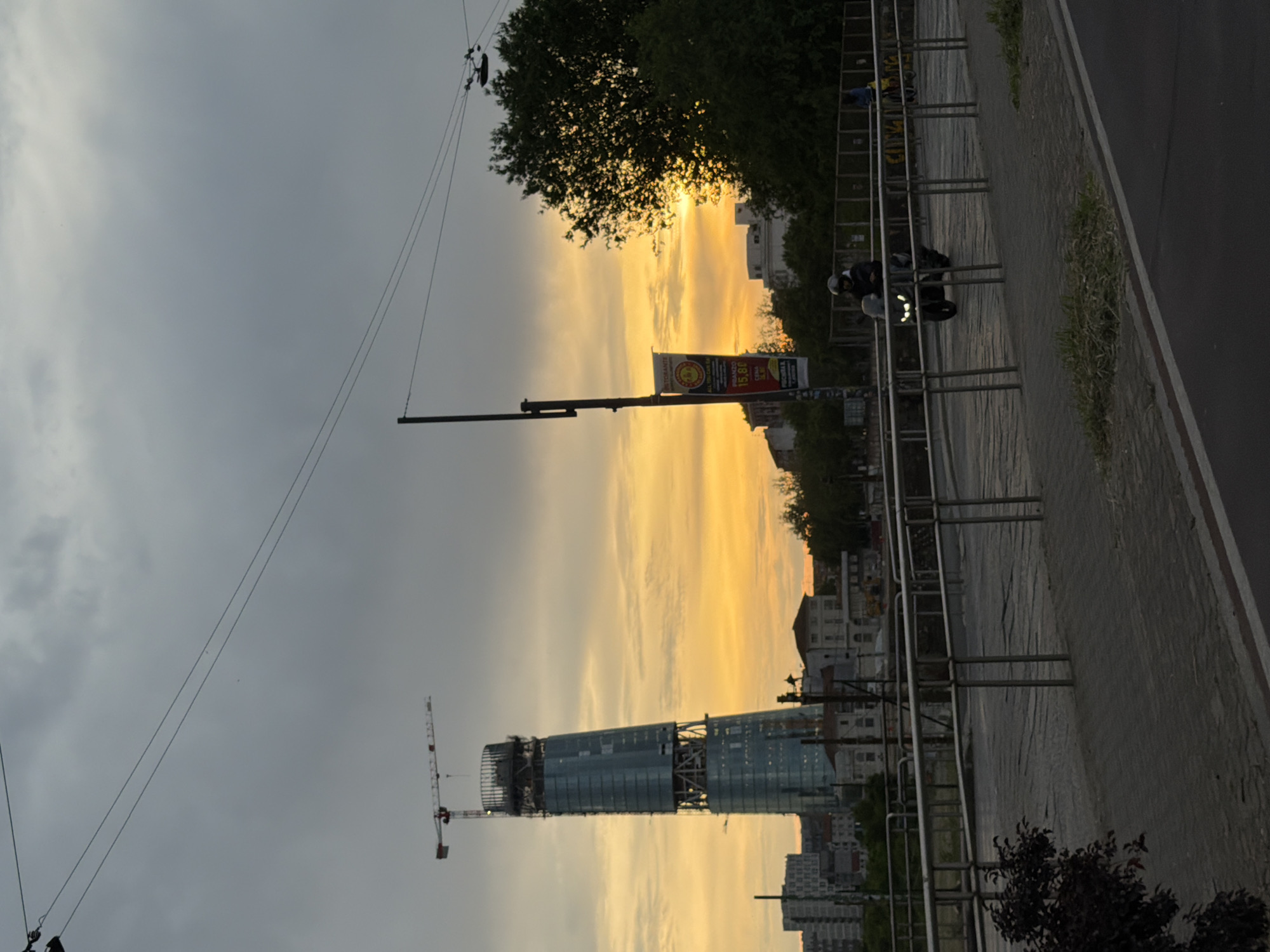 The image depicts a street scene in Milan, Italy, at dusk or dawn, as evidenced by the orange and yellow sky. Prominently featured is a high-rise building under construction, with a crane visible next to it. Utility poles and wires stretch across the sky. A street, with a sidewalk lined by metal railings or bike racks, runs along the right edge of the image. A person on a motorcycle is partially visible on the sidewalk. Various other buildings can be seen in the distance, contributing to the urban landscape. A large advertising banner is also attached to a pole. The overall scene is muted in color, and the street appears to be relatively quiet.