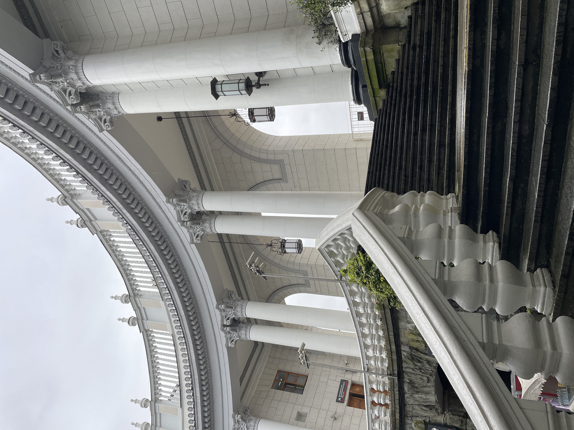 The image shows a section of the Sochi Train Station in Russia. The architectural style features a colonnade with white columns and ornate detailing. Black wrought iron stairs are visible on the right. Lighting fixtures hang from the ceiling under the colonnade. A portion of the building’s exterior is visible in the background. No people are visible in the frame.