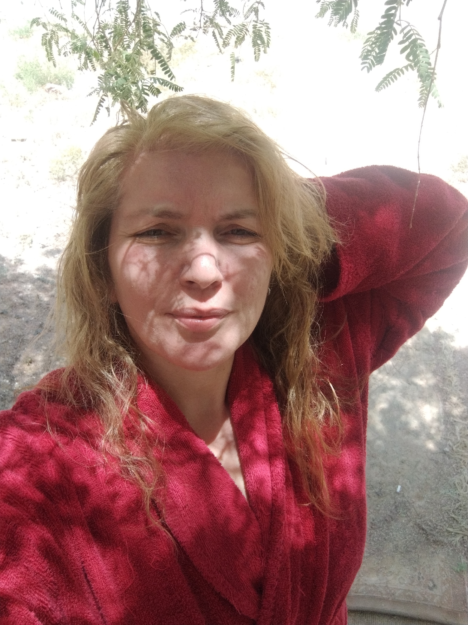 A woman is depicted in a selfie, wearing a red robe. The sunlight filters through foliage, casting shadows across her face. The background is slightly out of focus, suggesting an outdoor setting. The location is Al Mas'oudi, United Arab Emirates.