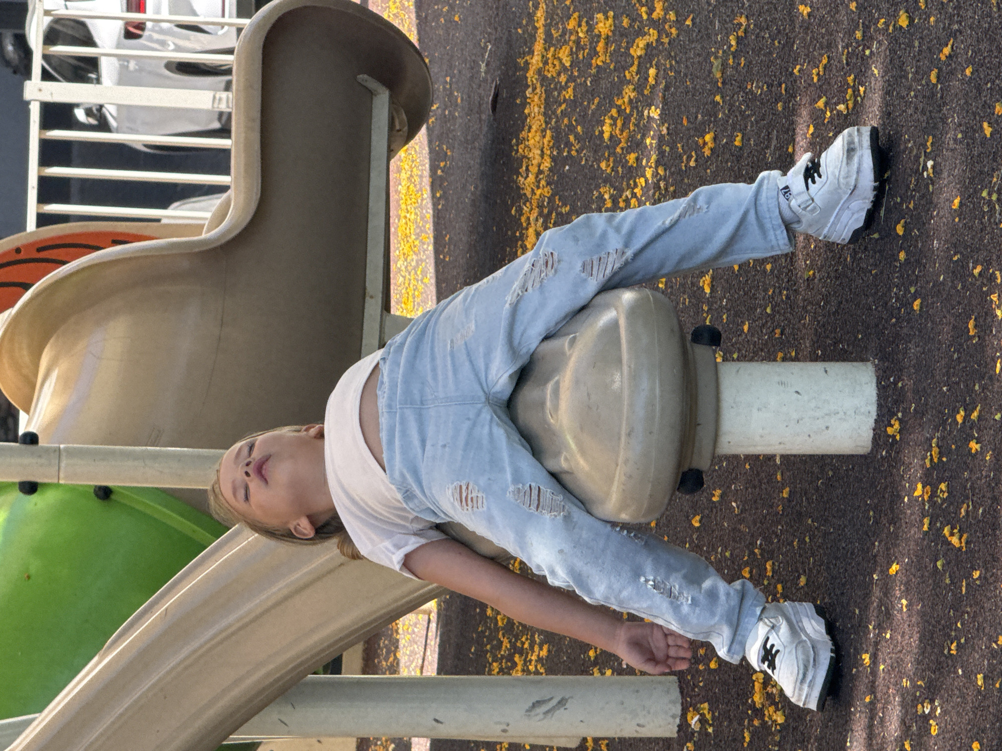 A young child with blonde hair is sleeping on a playground slide in the city of Unknown, Unknown. The child is wearing a white t-shirt, ripped light blue jeans, and white platform sneakers. The child's head is tilted back and their eyes are closed. The slide is a light brown color and is part of a larger playground structure that includes a green section and metal railings. The ground around the slide is covered in dark brown material with scattered yellow petals. In the background, a white SUV is partially visible behind the railings. The lighting suggests it is daytime.