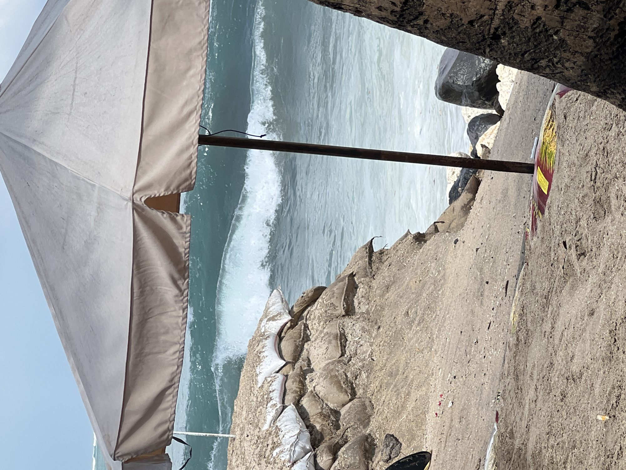 The image shows a beach scene in Kuta, Indonesia, with a large beach umbrella in the foreground. The ocean waves are breaking in the background, and there are sandbags stacked on the left side of the image, indicating erosion control measures. A surfboard with colorful designs is lying on the sand near the umbrella pole. The beach appears to be made of sand and some scattered debris. A palm tree trunk is visible on the right side of the frame. The sky is blue with some scattered clouds.